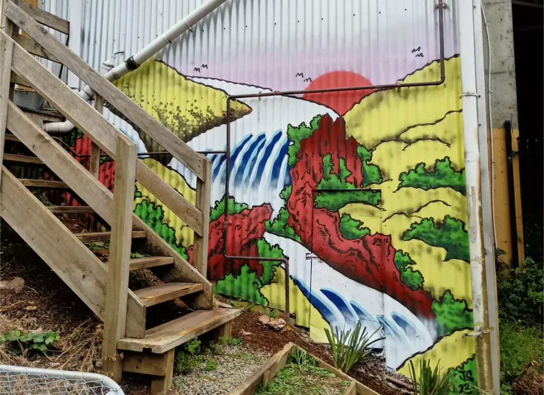 Colorful mural of a mountain landscape with a waterfall, green vegetation, red cliffs, a yellow mountain, and a red sun on a corrugated metal wall, with a wooden staircase in front.