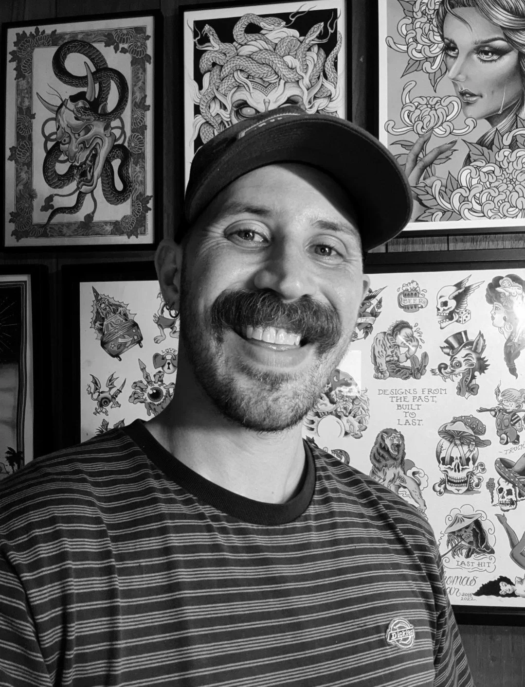 A man with a beard and mustache smiling, wearing a striped shirt and a cap, standing in front of wall art featuring various tattoo-style designs including a dragon, a snake, a woman, and other artistic illustrations.