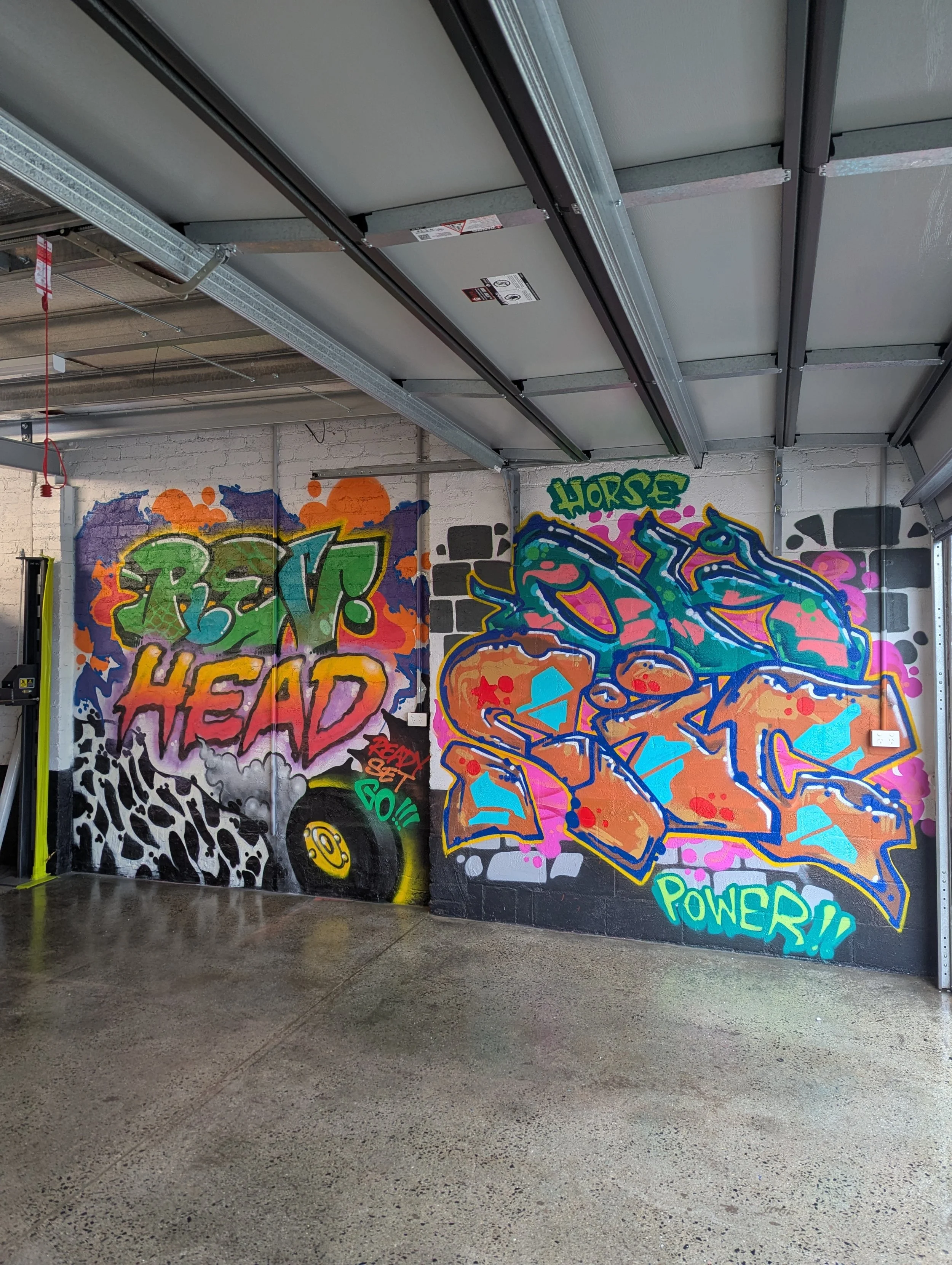 Indoor garage with concrete floor, graffiti-style artwork painted on the walls, including words like 'REV!', 'HEAD', 'POWER!', and various colorful designs.