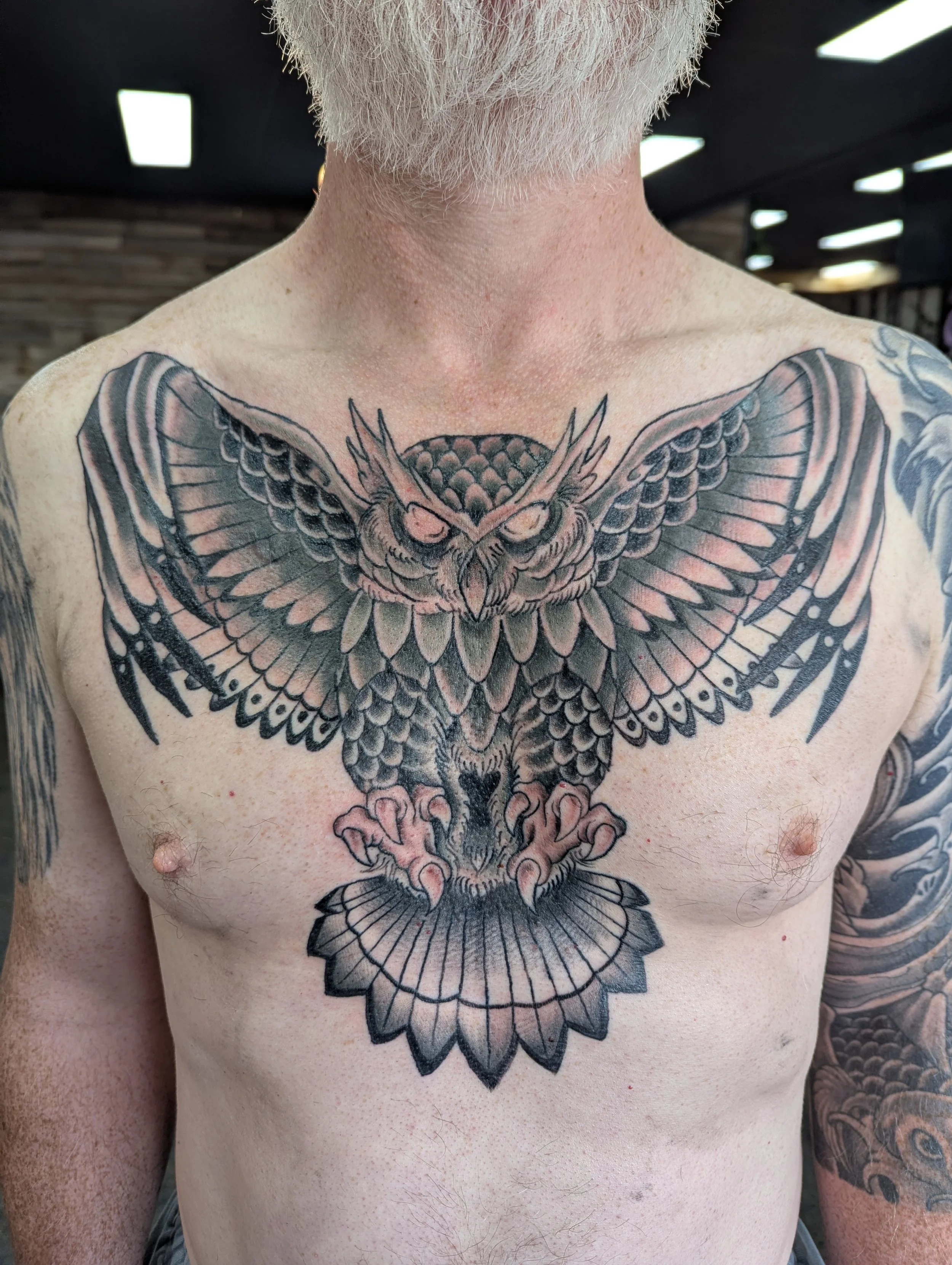 A person's chest and upper torso with a large, detailed black and grey owl tattoo on the chest and shoulders, partially visible arm tattoos, light skin, and white facial hair.
