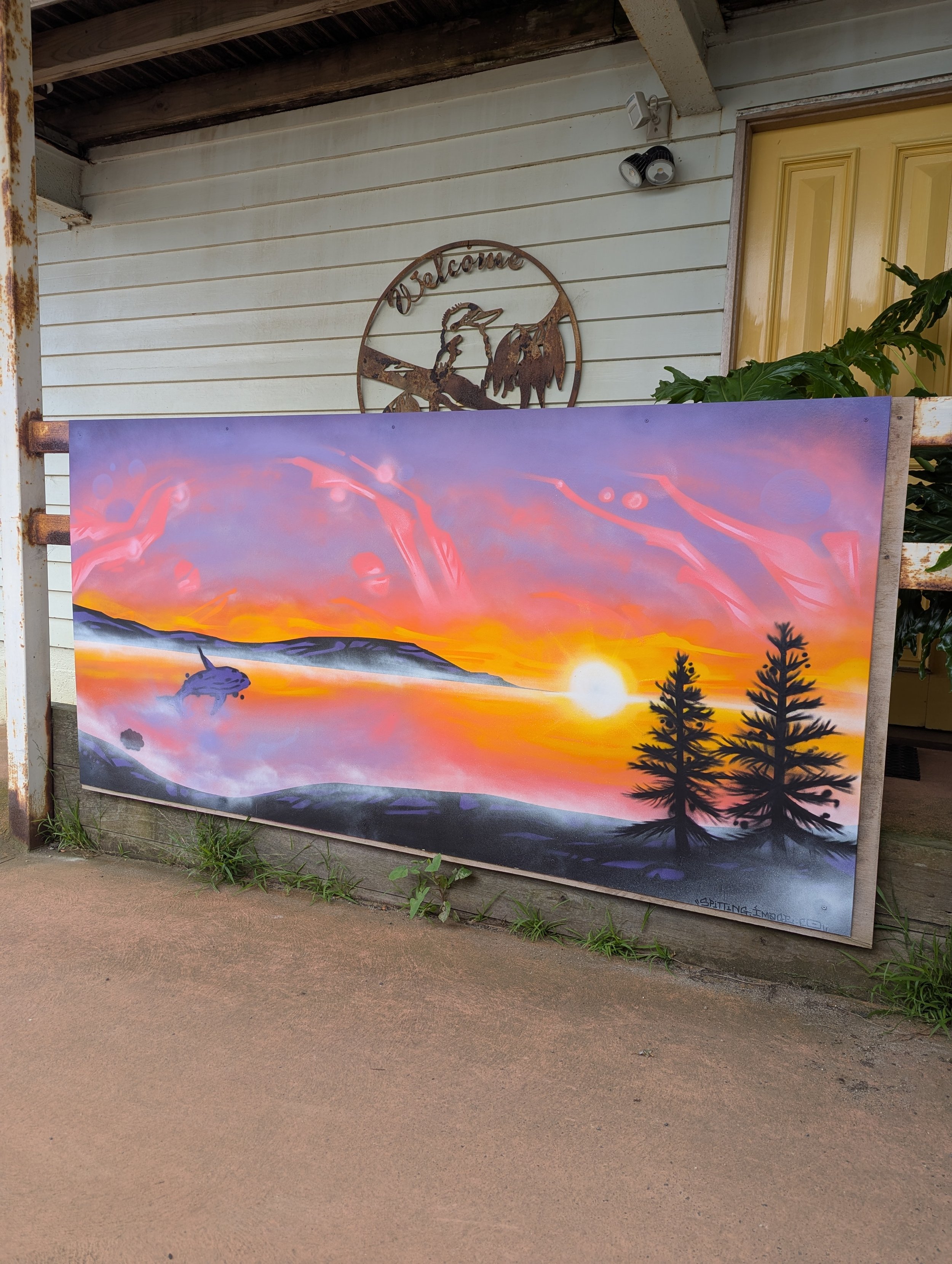 A sunset landscape painting with a vibrant sky of pink, orange, and purple hues, silhouettes of pine trees, and rolling hills in the distance, displayed outdoors on a wooden fence.