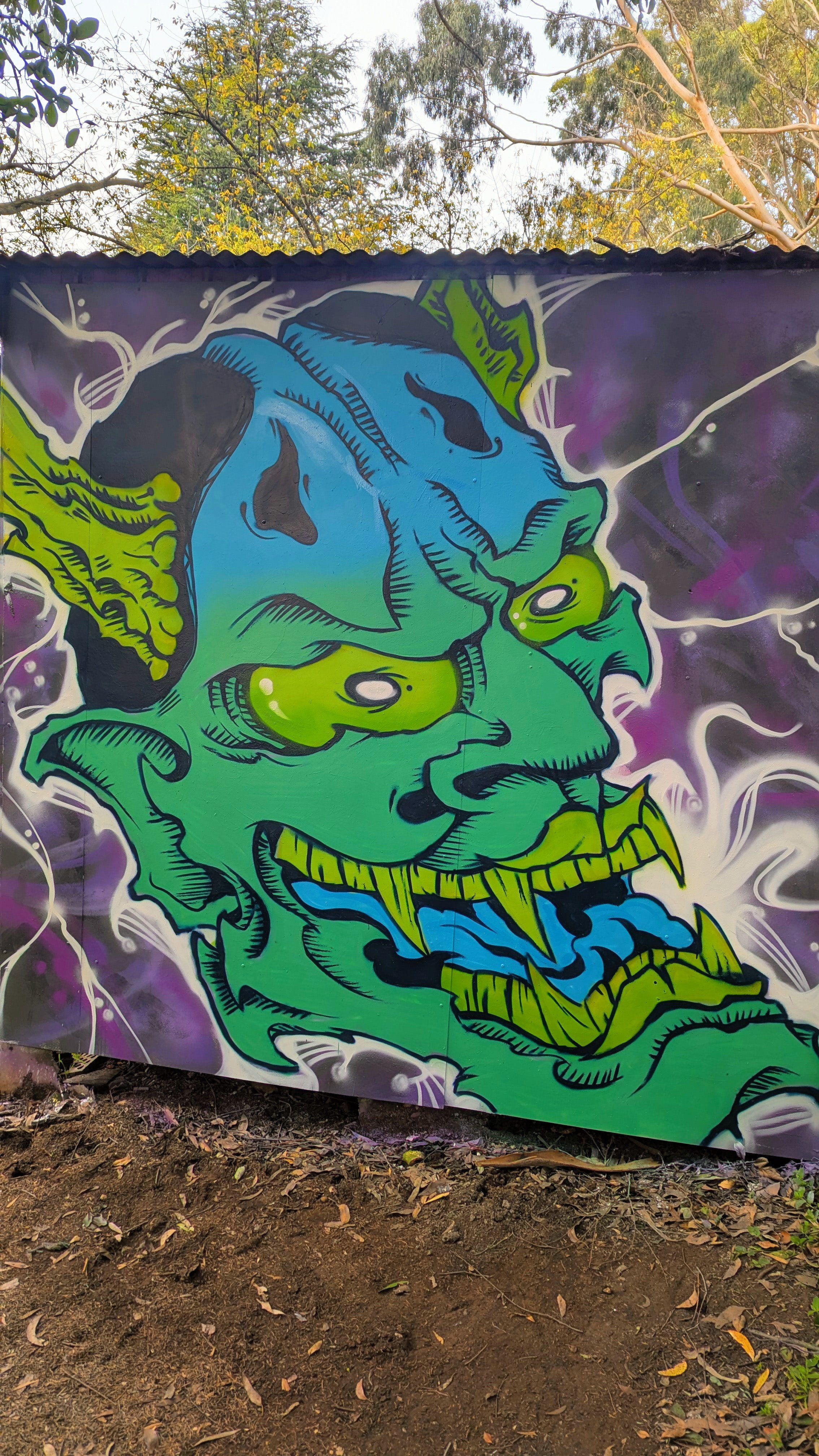 Colorful graffiti mural of a monstrous creature with a blue face, green body, black eyes, and sharp yellow teeth, set against a dark background with purple and white accents.