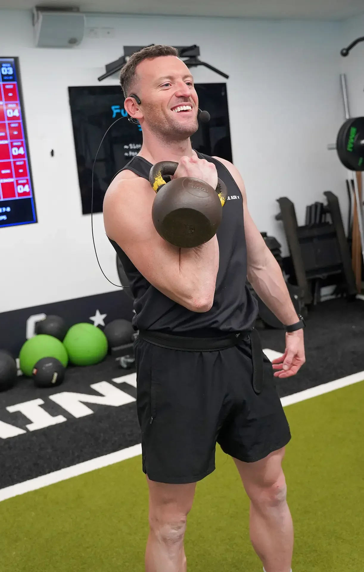 A man with a headset and a microphone is smiling while performing a kettlebell exercise in a gym. He is holding a kettlebell in his right hand across his shoulder. The gym has various exercise balls on the floor and fitness equipment against the wall.