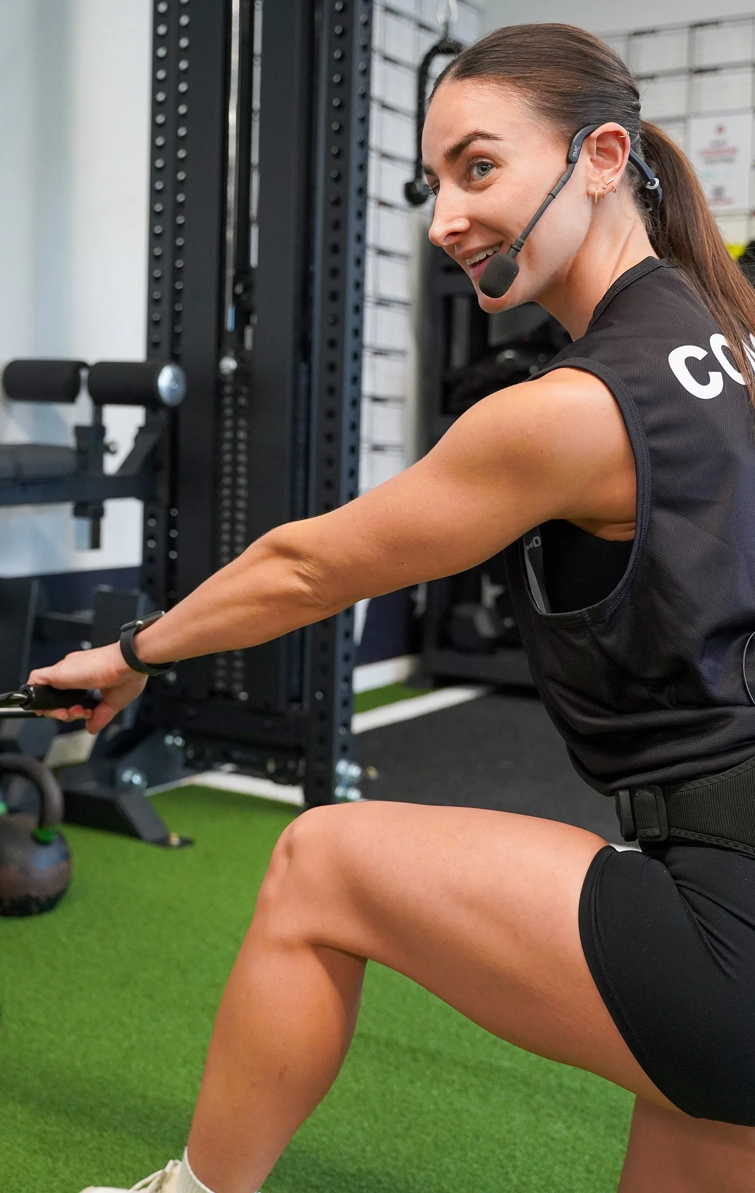 A woman in workout attire using a resistance band at a gym, wearing a headset and smiling.
