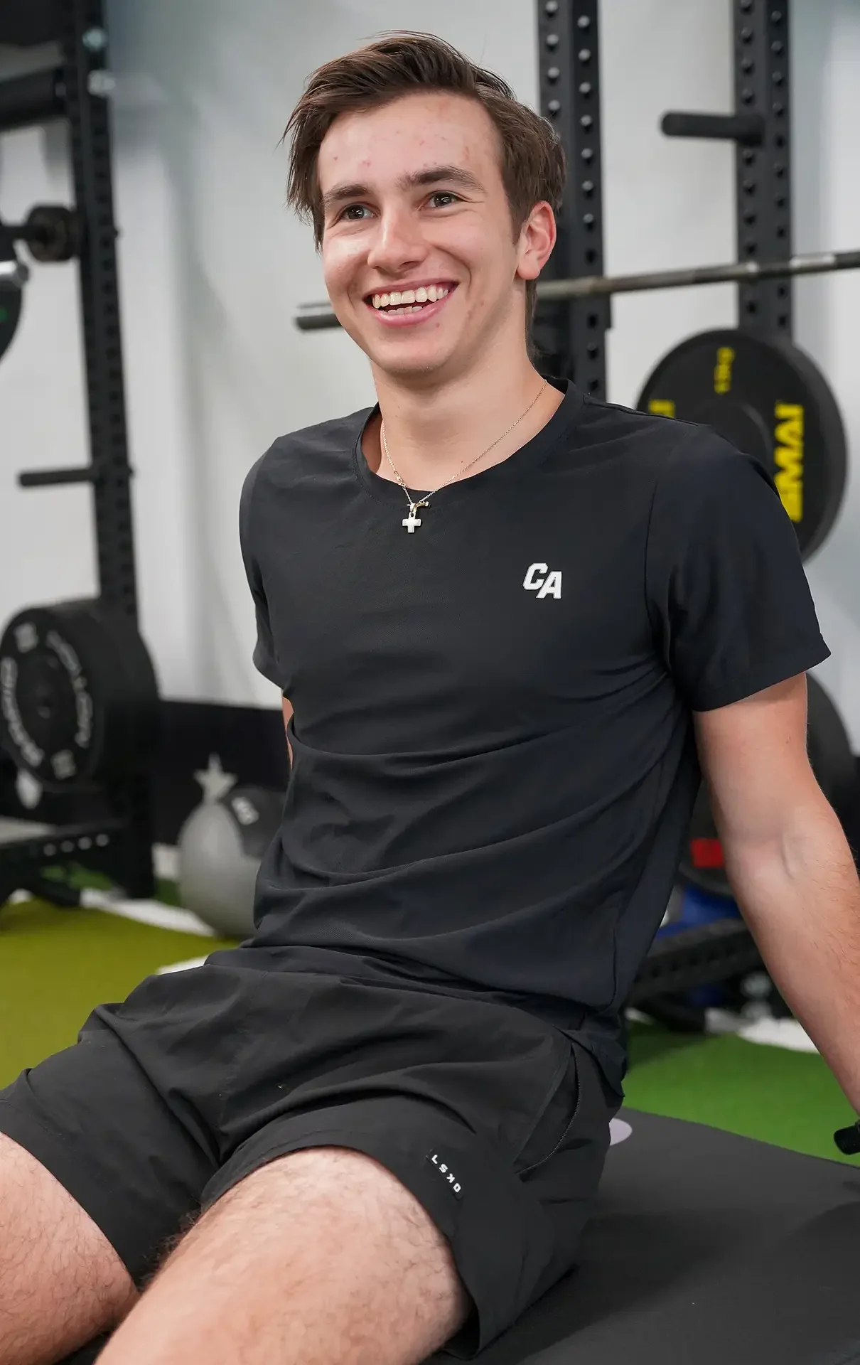 A young man smiling and sitting on a workout mat in a gym, surrounded by exercise equipment including weight plates and barbells.