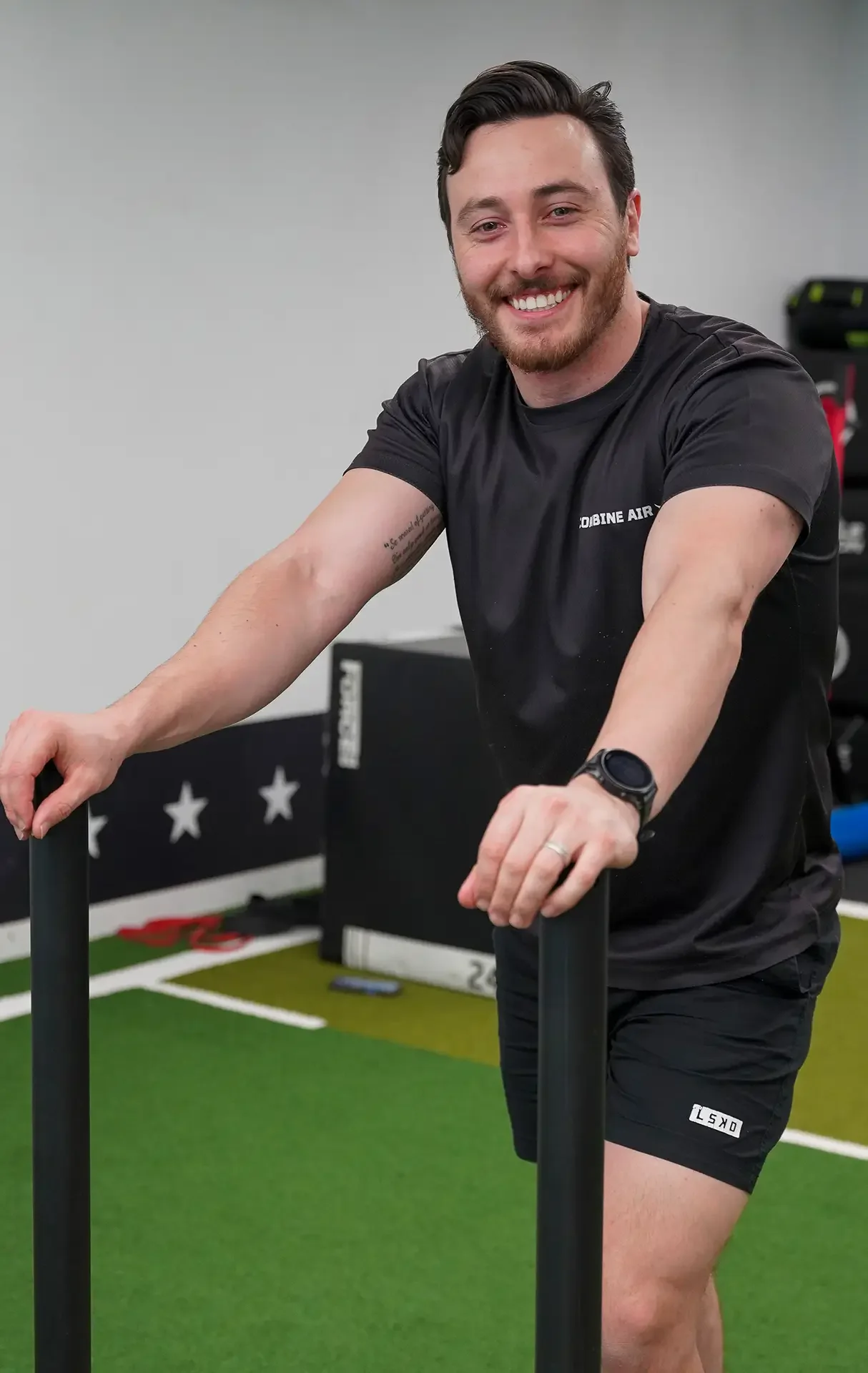 Man in black sportswear smiling, standing indoors on green turf, holding black bars, with gym equipment in the background.