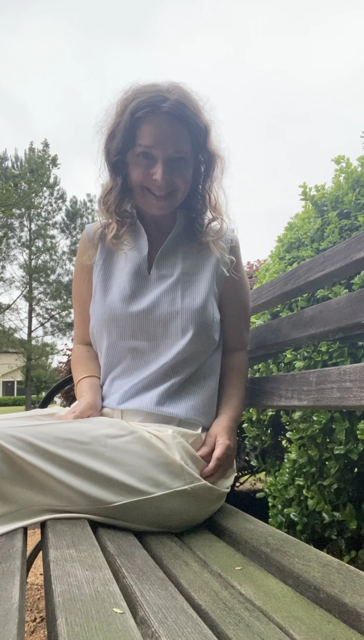 A woman with curly hair sitting on a wooden park bench, smiling, surrounded by green bushes and trees, on an overcast day.