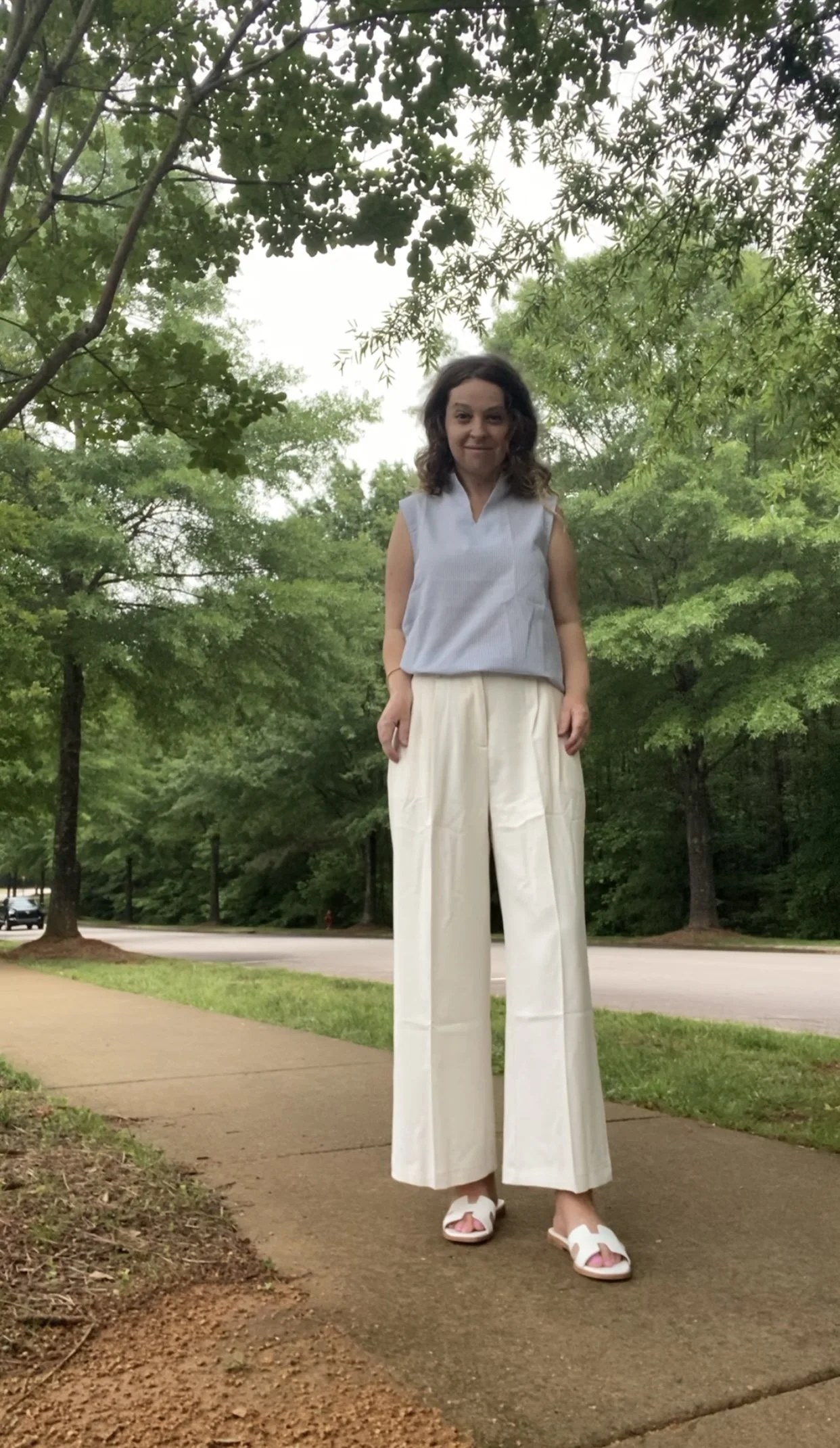 A woman standing outdoors on a sidewalk with trees and greenery in the background. She is wearing a light blue sleeveless top, wide-legged white pants, and white sandals. She has curly brown hair.