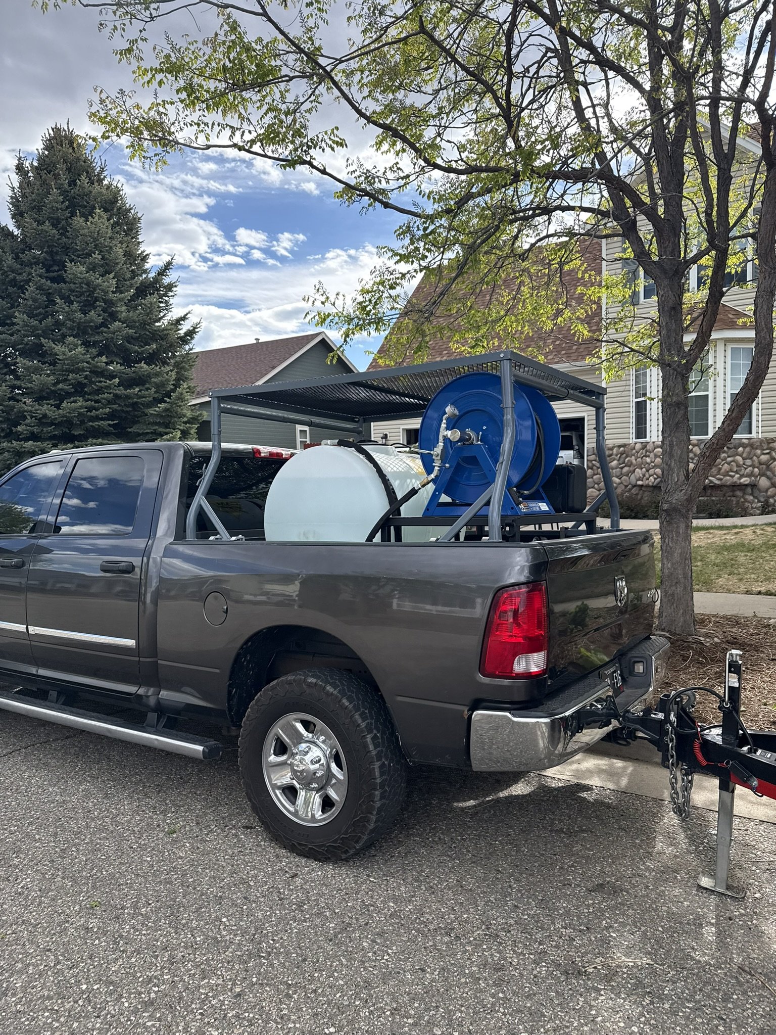 A gray pickup truck with a large water tank and hose reel mounted in its bed, parked on a residential street under a tree.