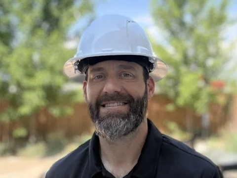 A man with a beard wearing a white hard hat and black work shirt, outdoors with trees and a fence in the background.