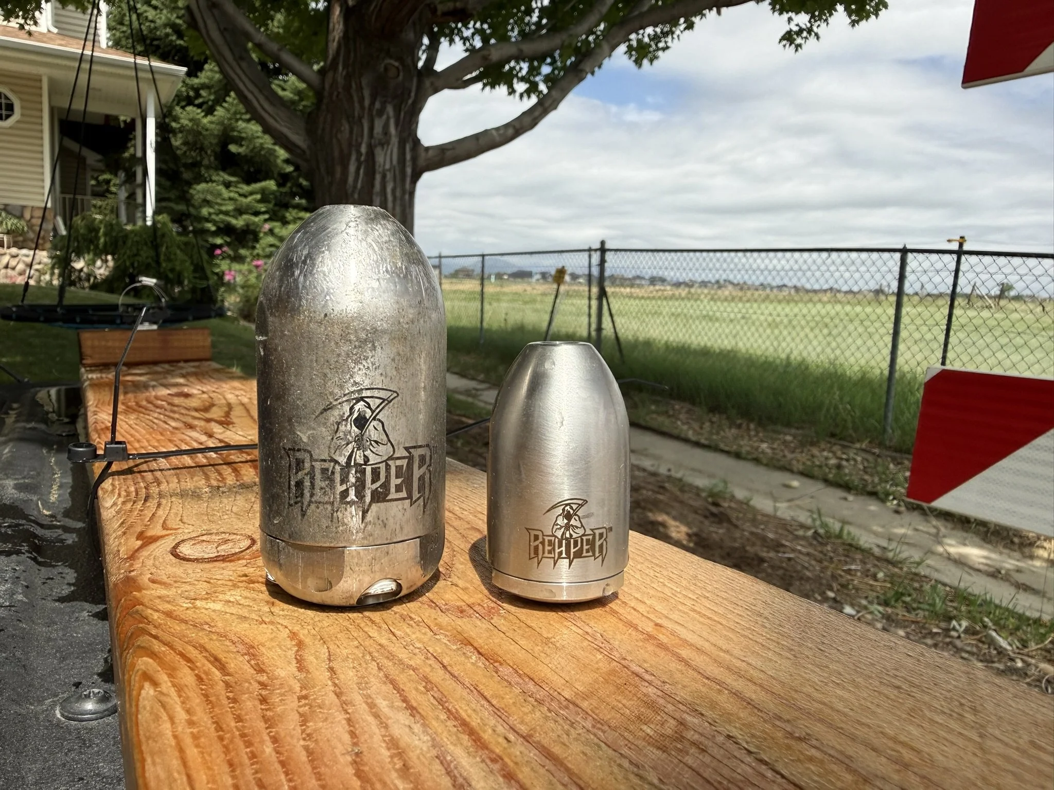 Close-up of two Reaper hydro Jet Drain cleaning nozzles sizes 1/4" and 3/8", placed on a wooden surface outdoors, with trees and a fence in the background.