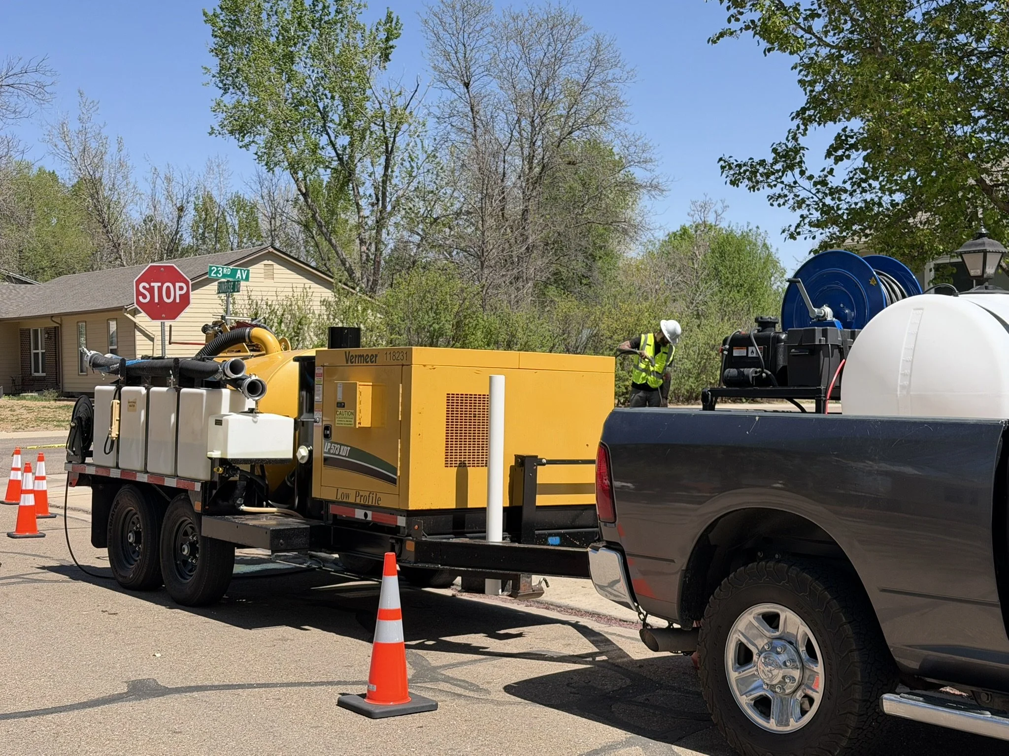 Utility workers  working on a slot trench for an electrical panel upgrade.  orange traffic cones around the worksite, and a man in a safety vest and hard hat is working near equipment on the street. A house and trees are visible in the background.