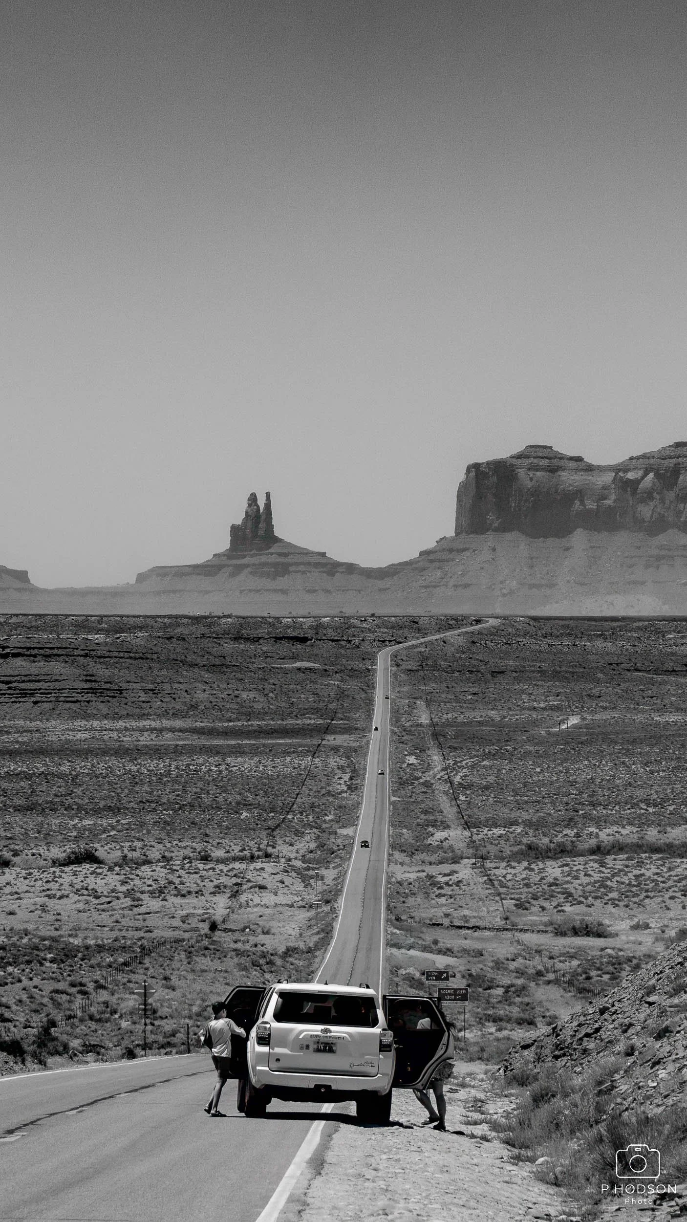 Black and white photo of a long, straight desert road stretching towards distant rock formations. People and a vehicle are at the roadside in the foreground.