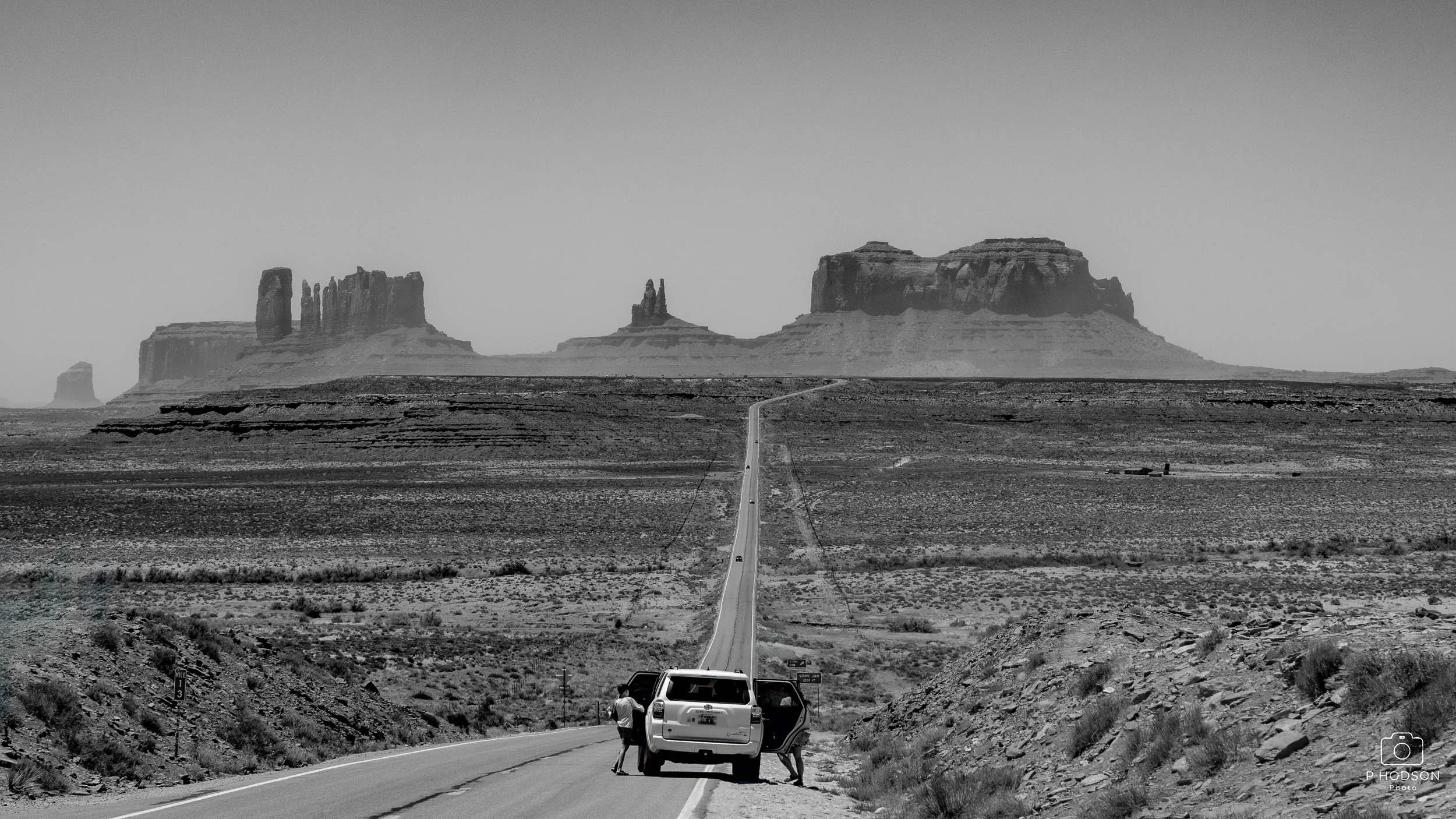 A black and white photo of a desert landscape with a long straight road leading towards distant rock formations, with cars and hikers in the foreground.