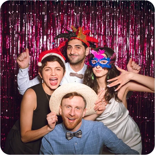 Group of four people celebrating, wearing festive and silly accessories, in front of a shiny pink backdrop.