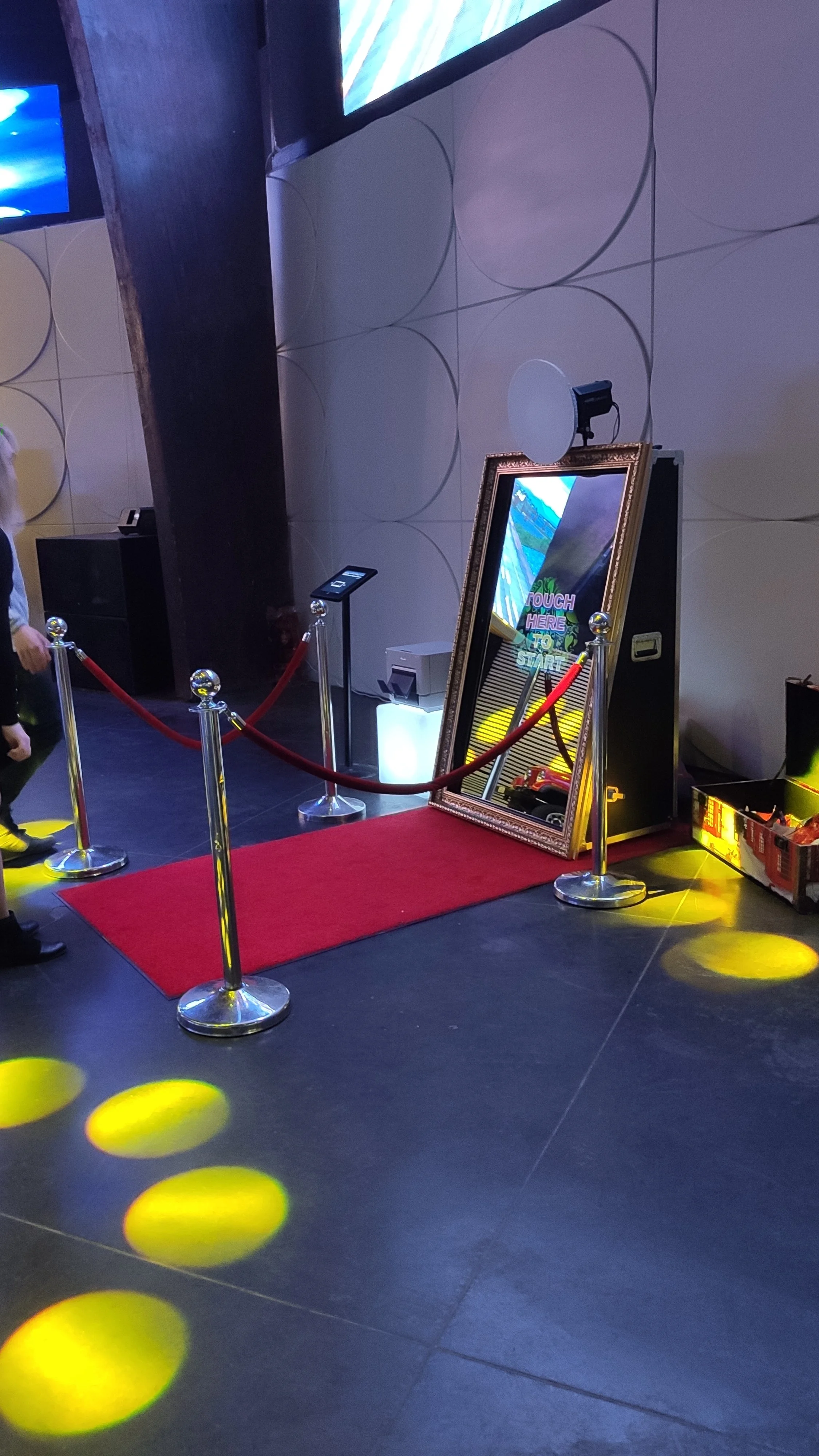 An indoor arcade or entertainment venue with a red carpet, a large framed mirror, and a mounted game or photo booth setup. The area is roped off with red velvet ropes and yellow circular floor lights.