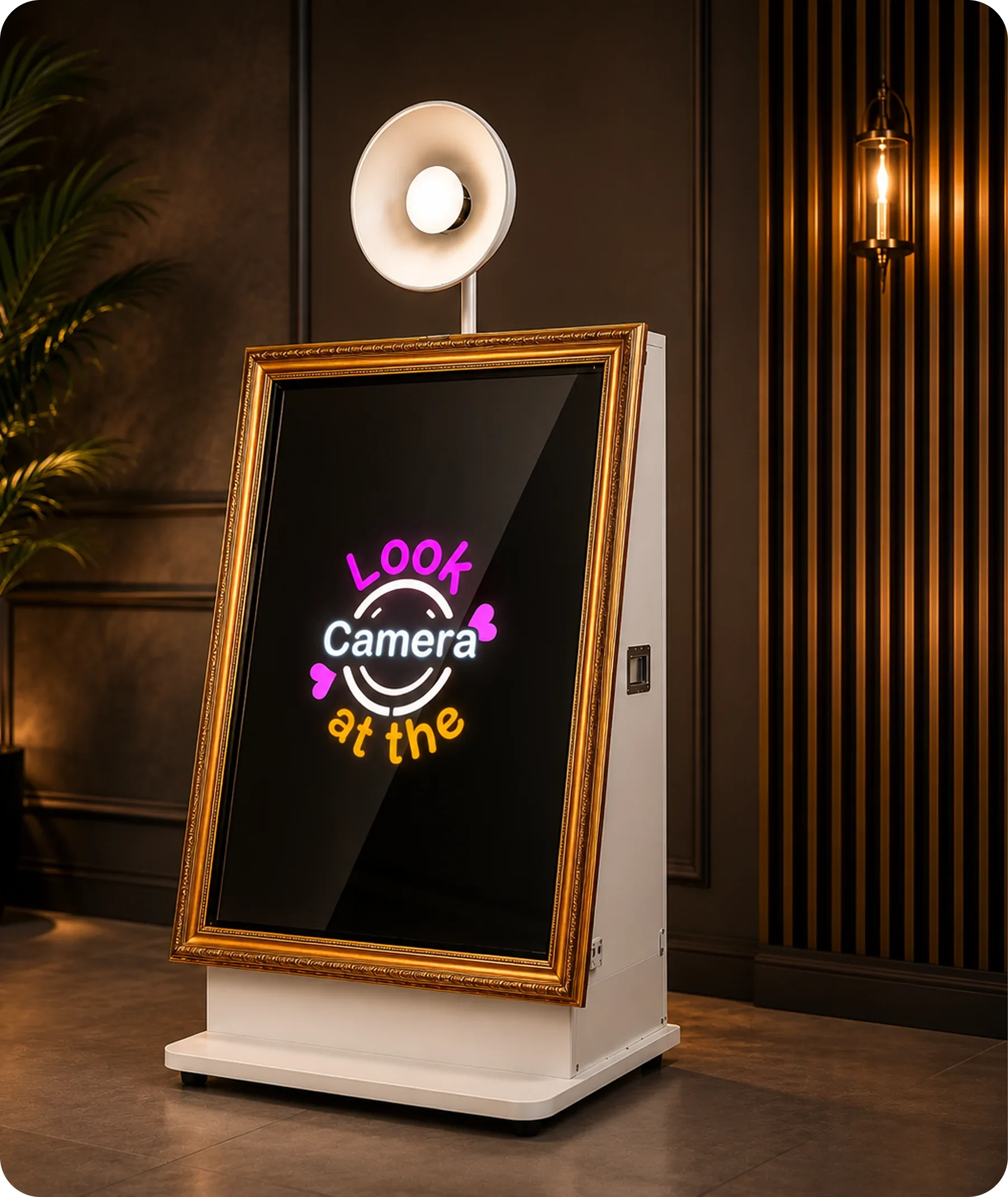 Photo of a photo booth with a gold frame, a digital screen displaying colorful text that says "LOOK at the camera," and a white stand. The booth is positioned indoors on a dark wood floor, with a wall featuring vertical gold and black stripes to the right and a plant to the left.