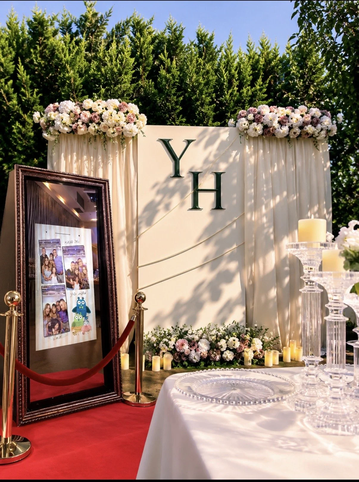 Decorative wedding or event photo booth setup with floral arrangements, candles, a framed photo display, and a backdrop with the initials "Y H" on a cream-colored background with drapes, set outdoors with greenery.