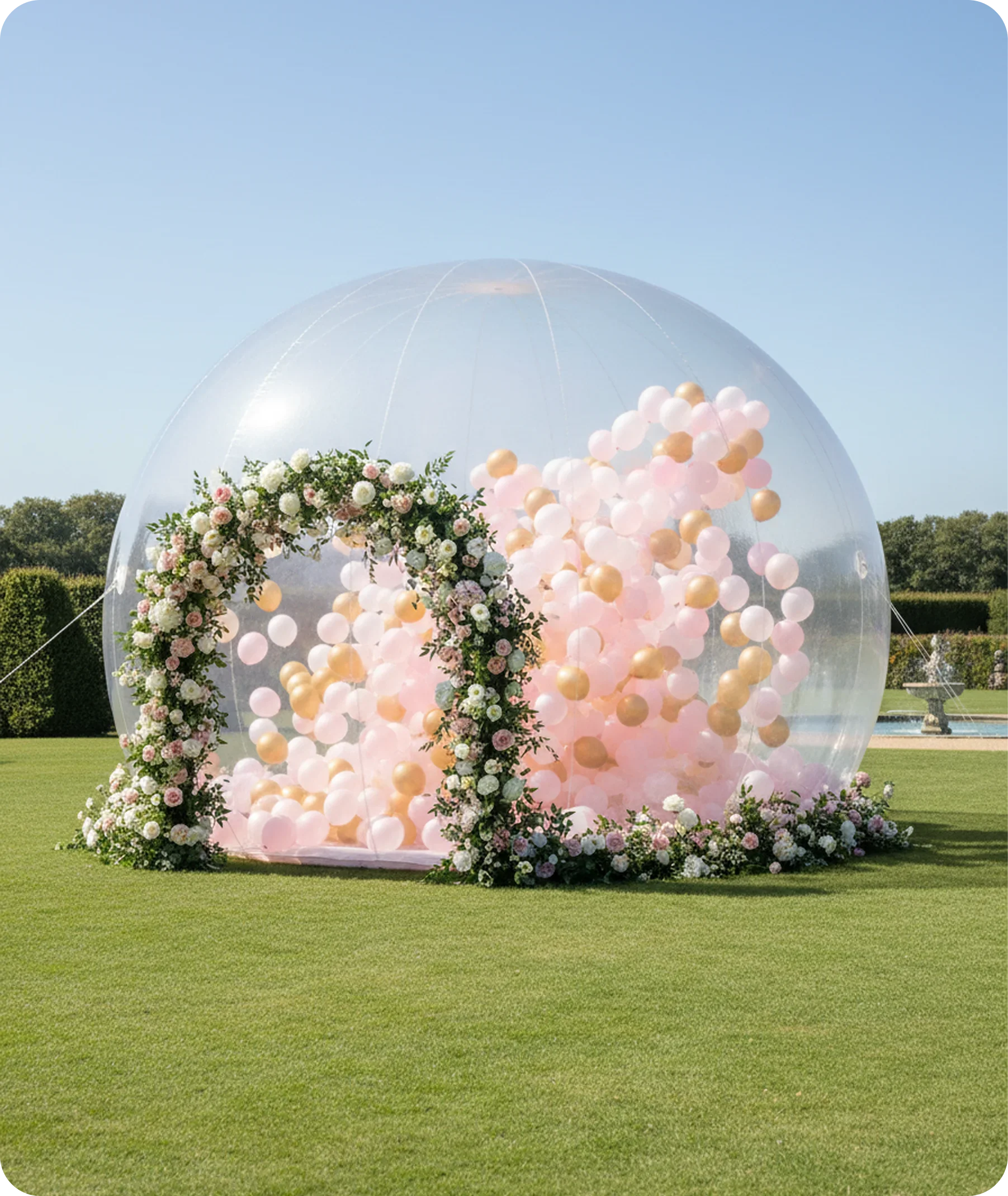 A large transparent dome surrounded by a floral arrangement of pink and white flowers on a grassy field with trees and a fountain in the background.