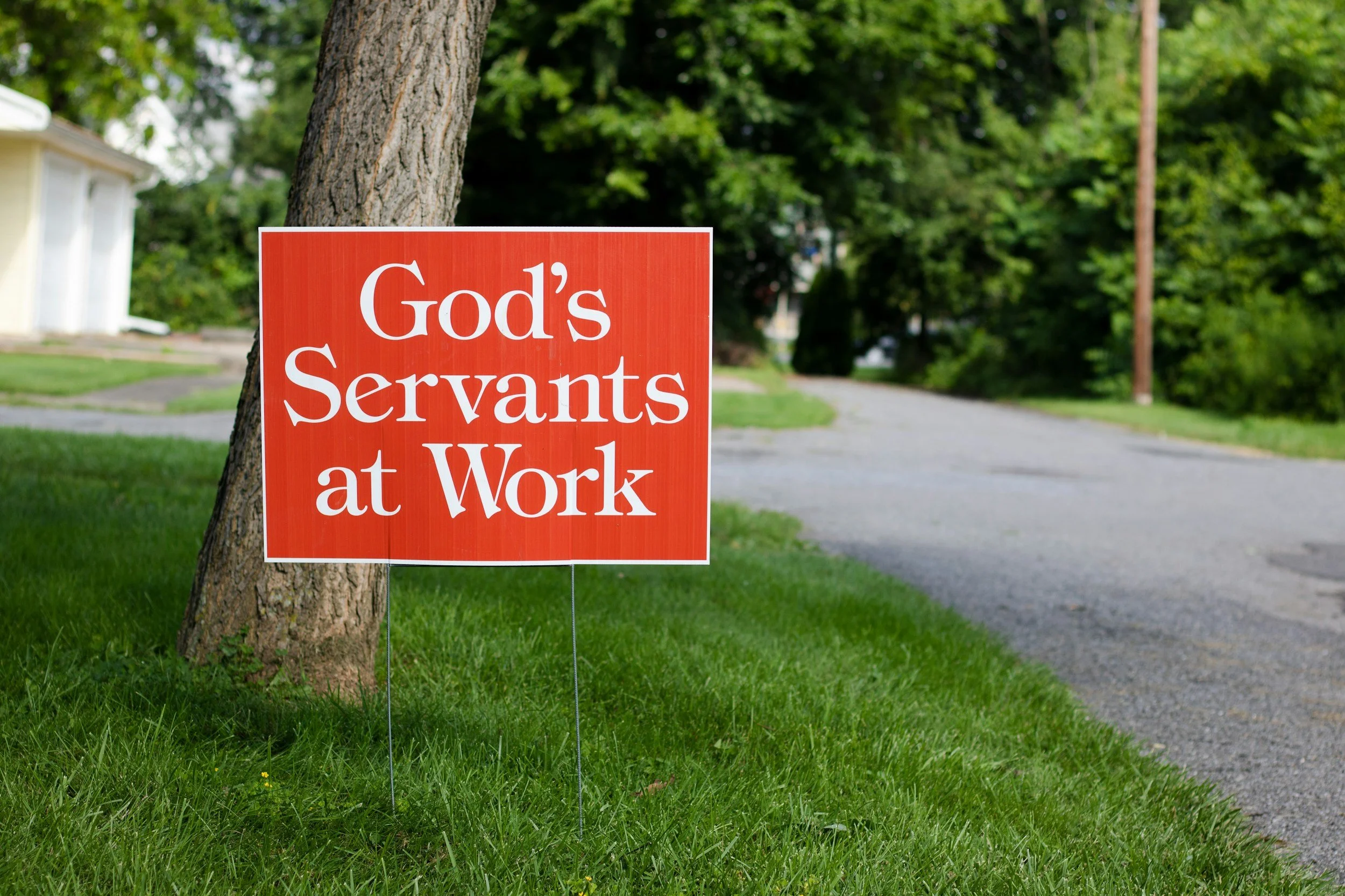 A red sign on a lawn next to a tree reads 'God's Servants at Work'. In the background, there is a driveway, some houses, and green trees.