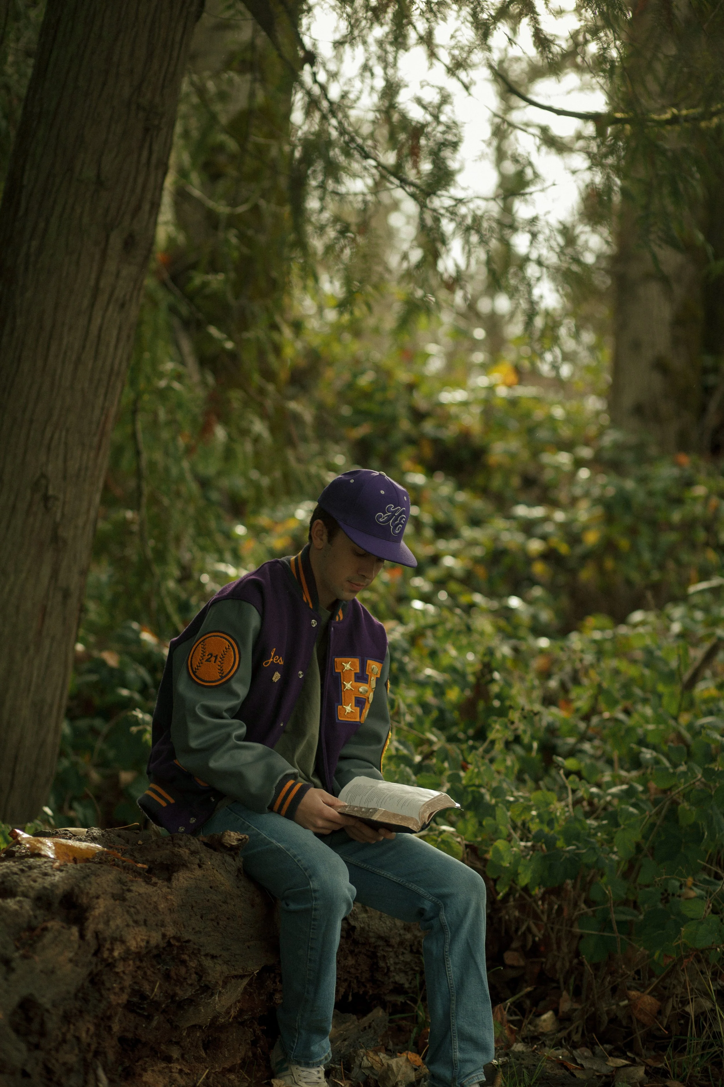 Young man sitting on a fallen log in a wooded area, reading a book, wearing a purple and green varsity jacket and a purple baseball cap.
