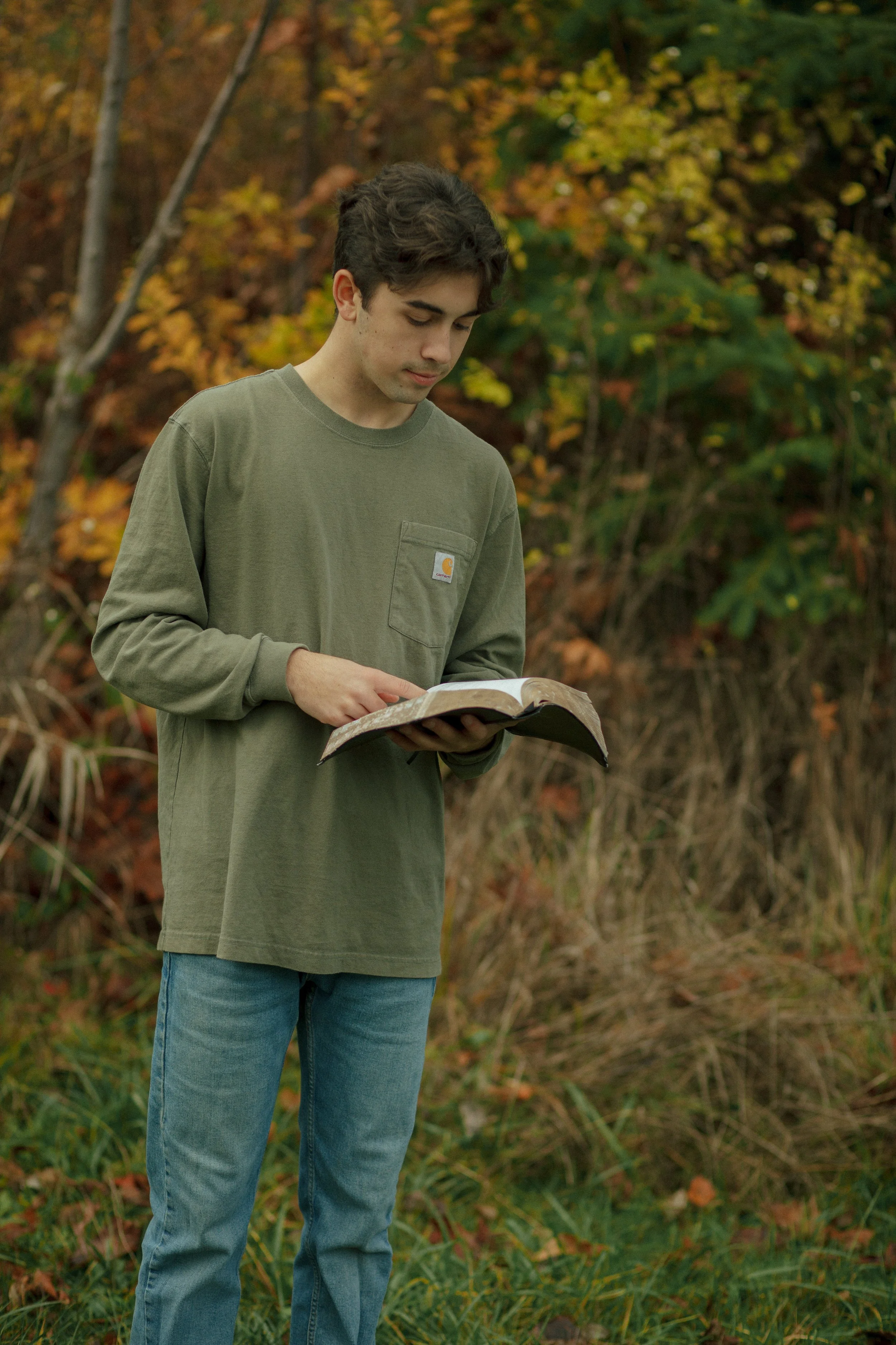 Young man wearing a green Carhartt shirt and blue jeans, standing outdoors in a wooded area during fall, looking down at a book he is holding.