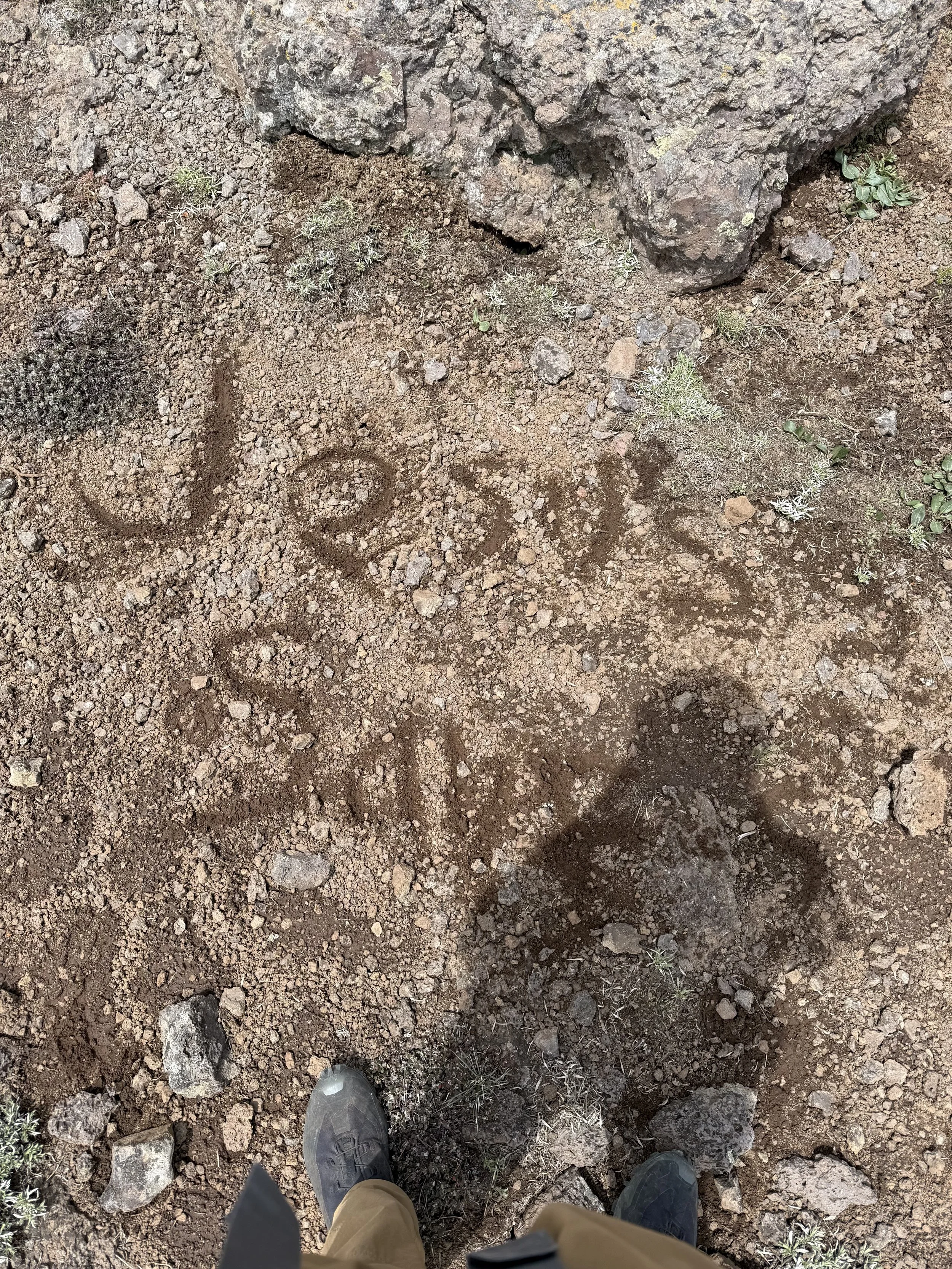 Photograph taken from a person's perspective standing on rocky, dirt ground with a few small plants; there are two people wearing hiking shoes, with one in gray shoes and the other in black shoes, visible at the bottom of the image.
