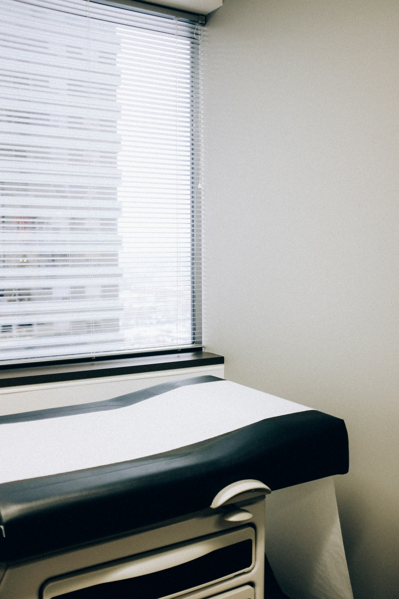 A hospital room with a window covered by white horizontal blinds. There is a white bed with black trim visible at the bottom of the image.