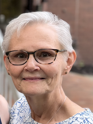 Close-up of an elderly woman with short white hair, glasses, and a blue-patterned top, smiling outdoors.
