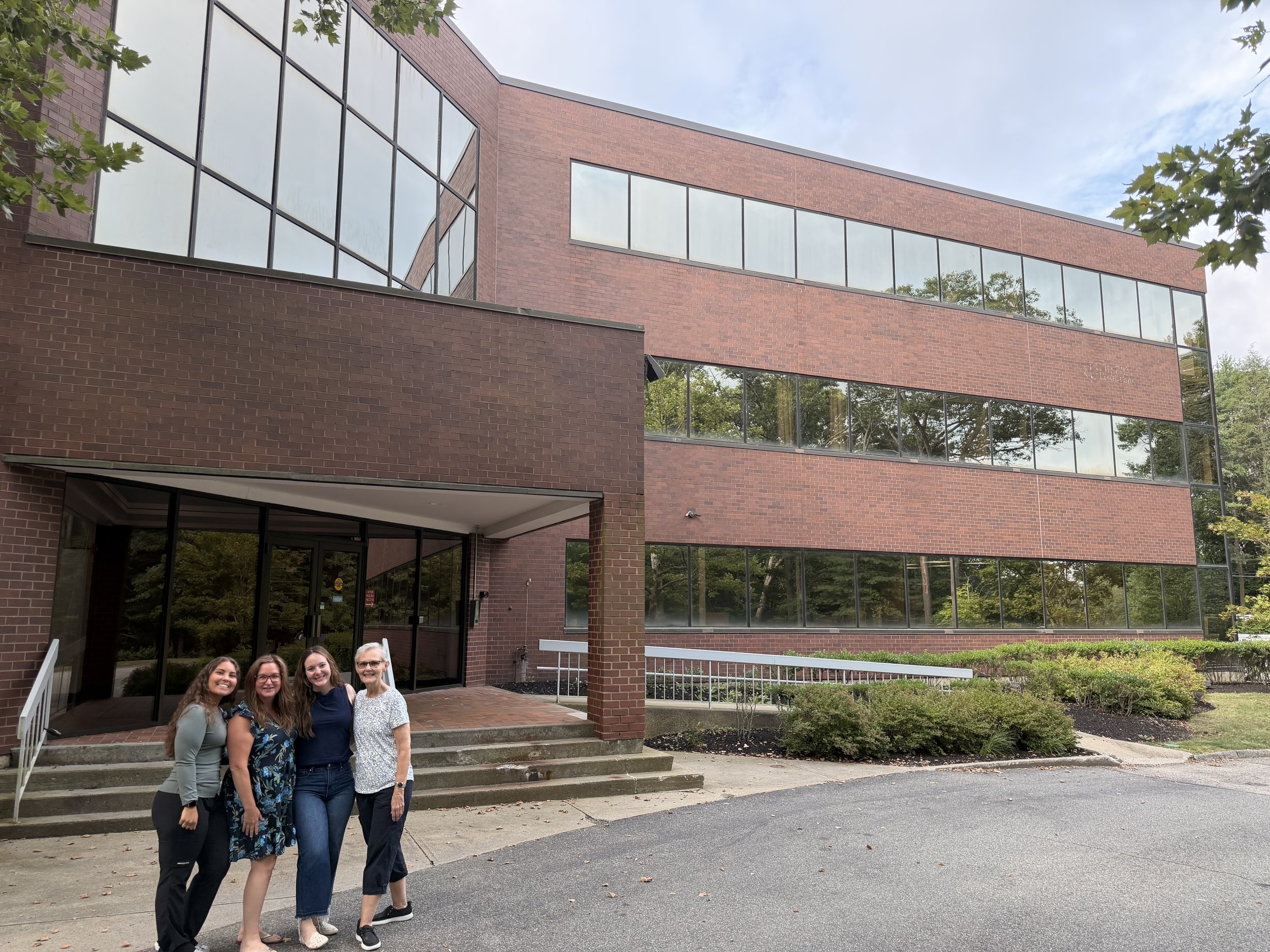 A group of five women standing on the sidewalk in front of a modern brick building with large glass windows, surrounded by greenery, on a cloudy day.