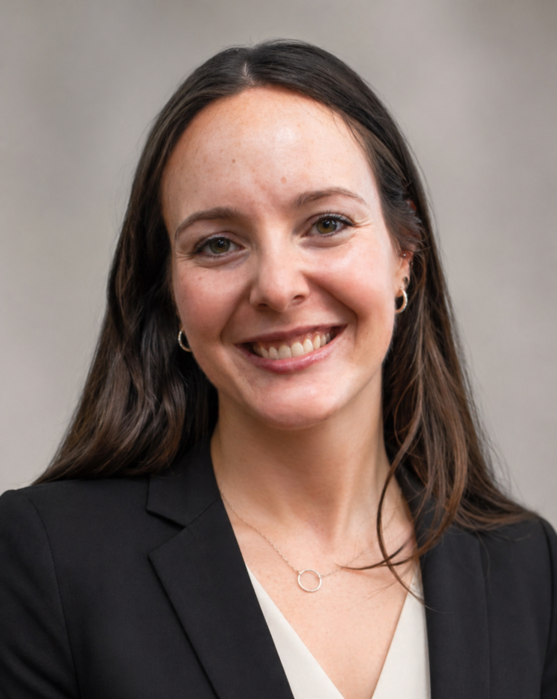 Close-up portrait of a woman with long brown hair, smiling, wearing a black blazer, white top, small hoop earrings, and a delicate silver necklace, standing against a neutral background.
