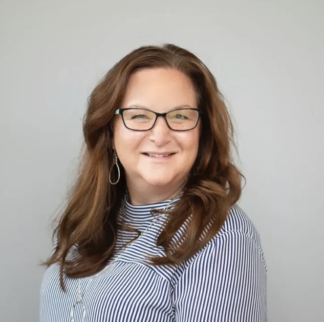 A woman with long brown hair, glasses, and earrings smiling at the camera, wearing a striped shirt, against a plain background.