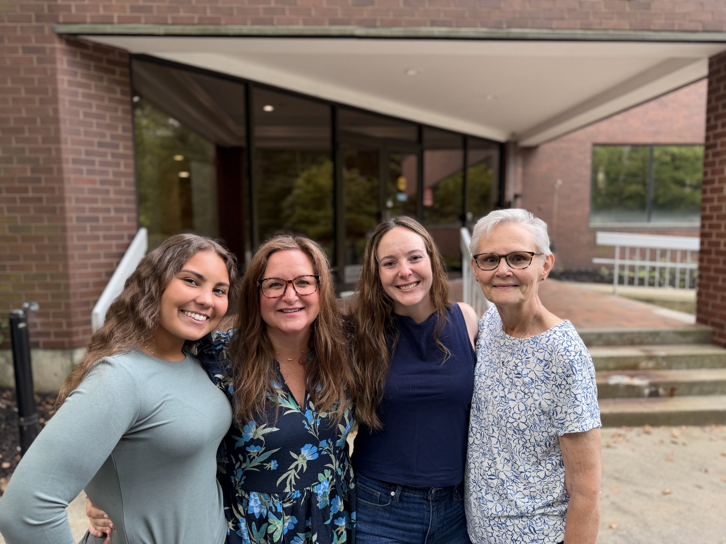 Four women standing arm in arm outside in front of a brick building, smiling for the camera.