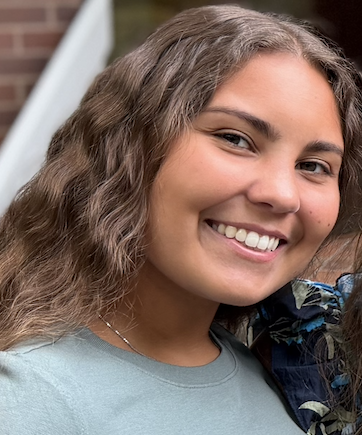 Close-up of a young woman with long, wavy brown hair smiling outdoors.