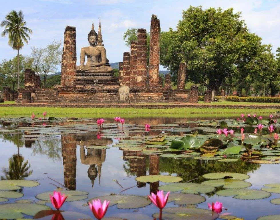 Thailand Travel. Ancient Buddha statue seated on a brick platform surrounded by old brick ruins. A pond with pink water lilies in the foreground, green trees, and a partly cloudy sky in the background. JadeInk Travel. Travel Advisor. Virginia