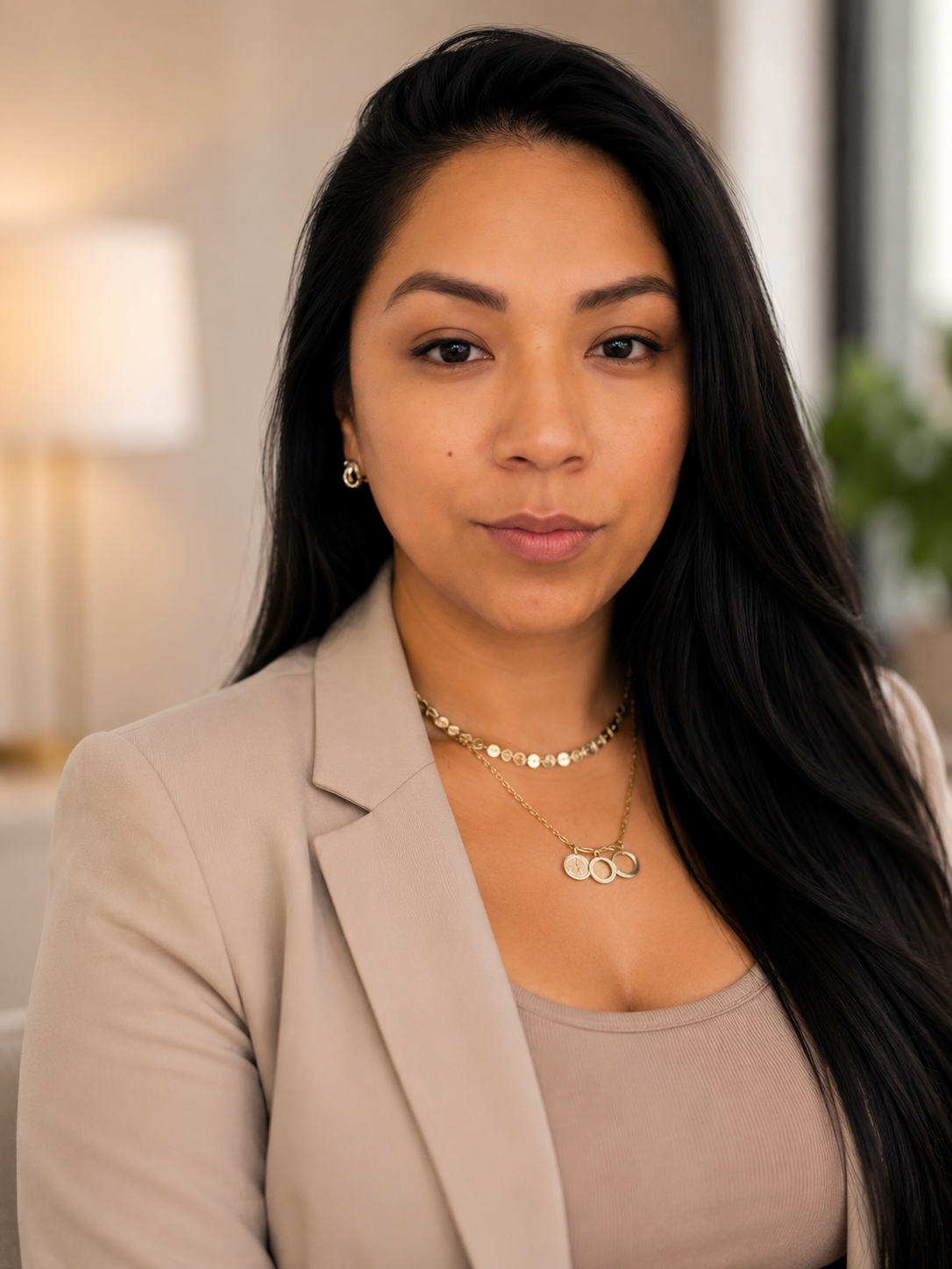 A woman with long black hair wearing a beige blazer, a beige top, and gold jewelry, including earrings, necklace, and a pendant with three circles, looking directly at the camera in a well-lit indoor setting.