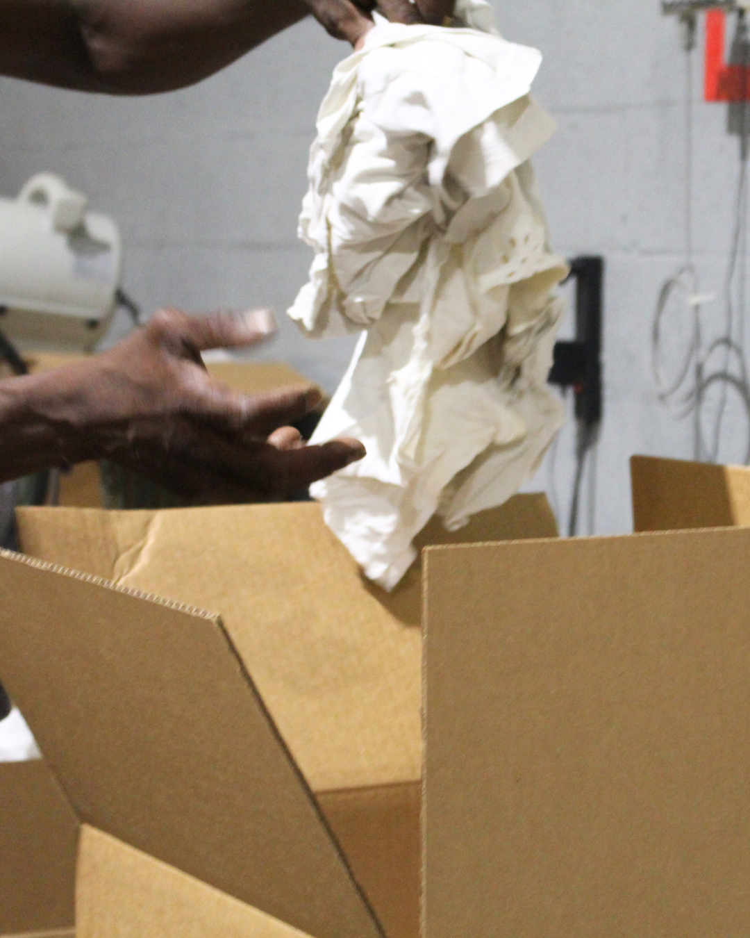 Hands placing white fabric wash cloths into a cardboard box in an industrial setting.