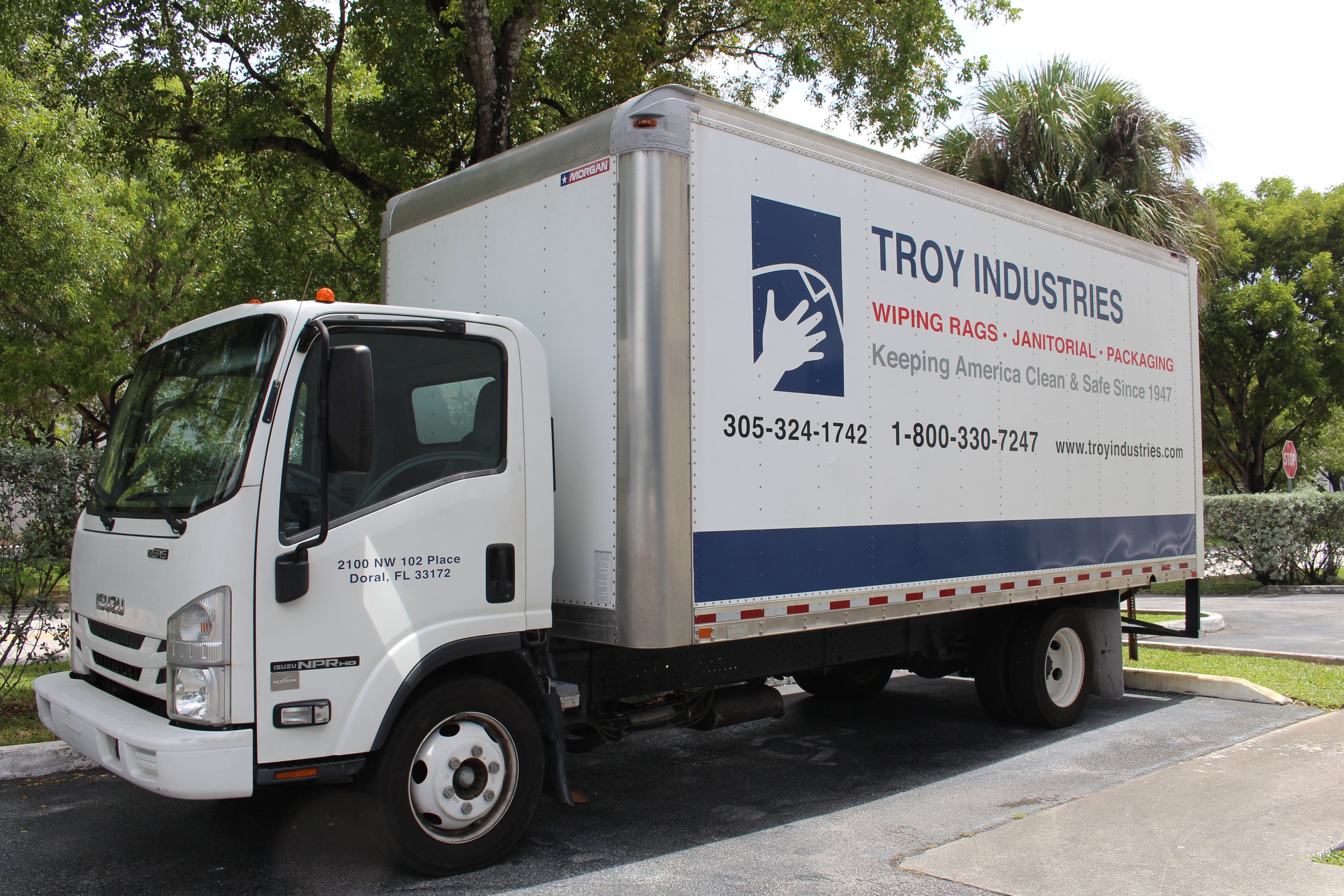 A white box truck with the logo of Troy Industries and text advertising wiping rags, janitorial supplies, and packaging. The truck is parked in a lot with trees and a stop sign visible in the background.