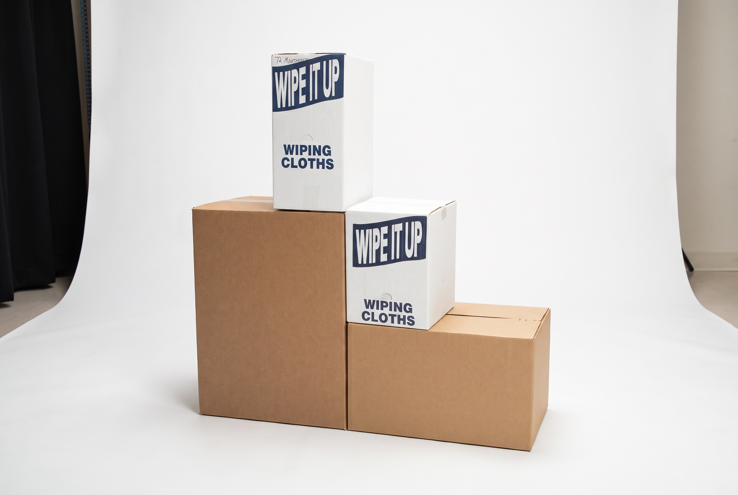 Three cardboard boxes arranged on a wooden surface against a white backdrop. Two white boxes are labeled "WIPING CLOTHS" with blue text and a blue rectangle, and one plain brown box.