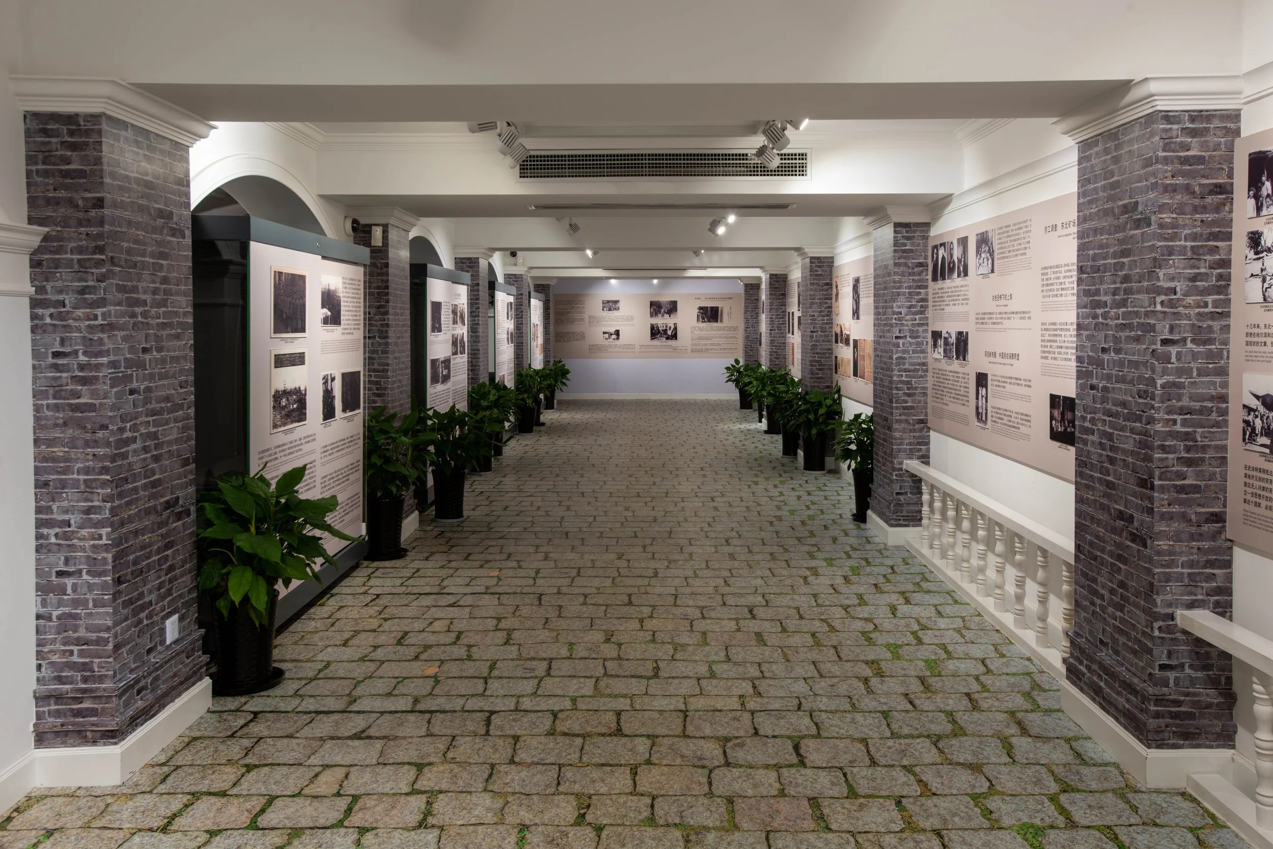 An indoor exhibition space with brick columns, white walls, and display panels featuring photographs and text, decorated with potted plants along the walkway.