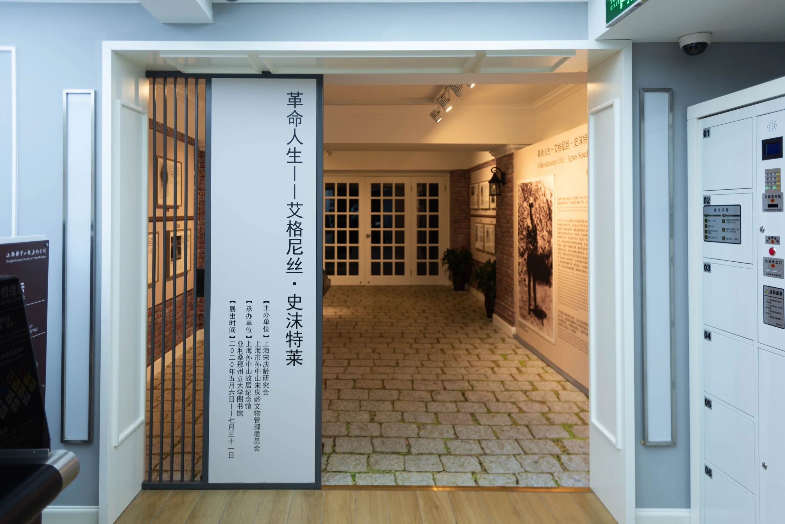 Entrance to an exhibition hall with a white gate, a tall white sign with Chinese and English text on the left, and lockers on the right. The background shows a hallway with brick walls and framed pictures, lit with warm lighting.
