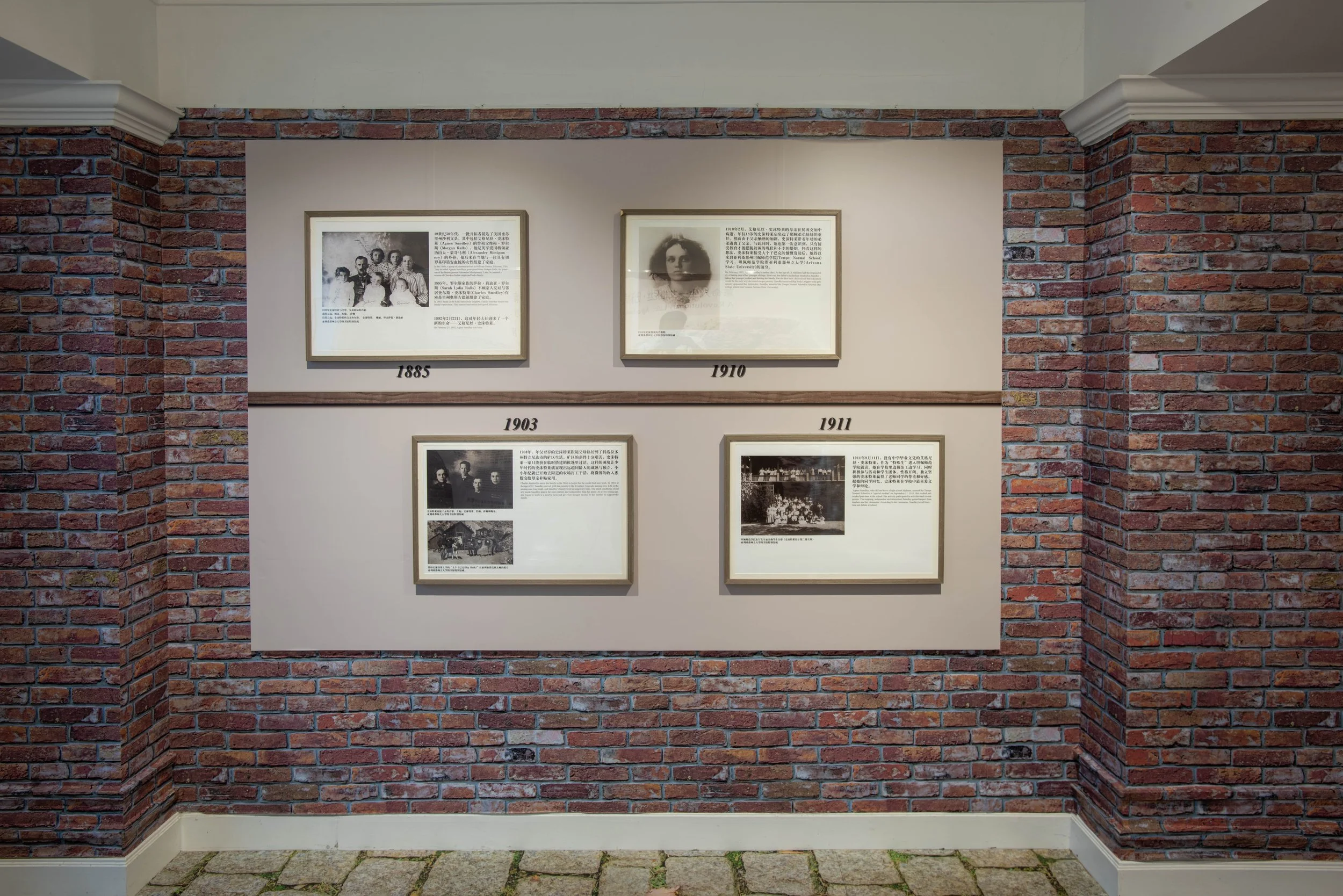 Exhibition wall with four framed black-and-white photographs and descriptions, surrounded by red brick walls.