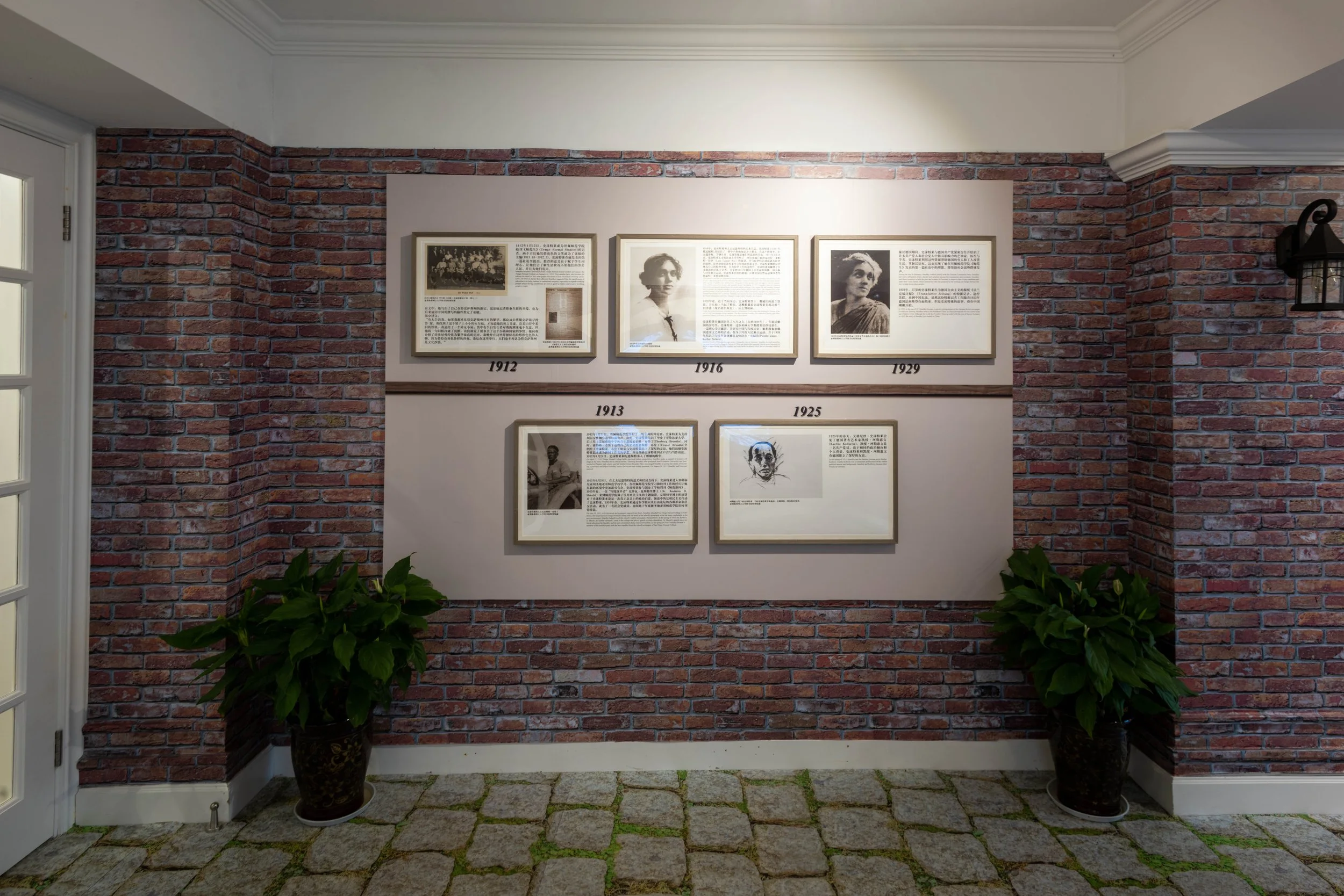 Exhibition wall with framed historical photographs and text panels, surrounded by brick walls and potted green plants.