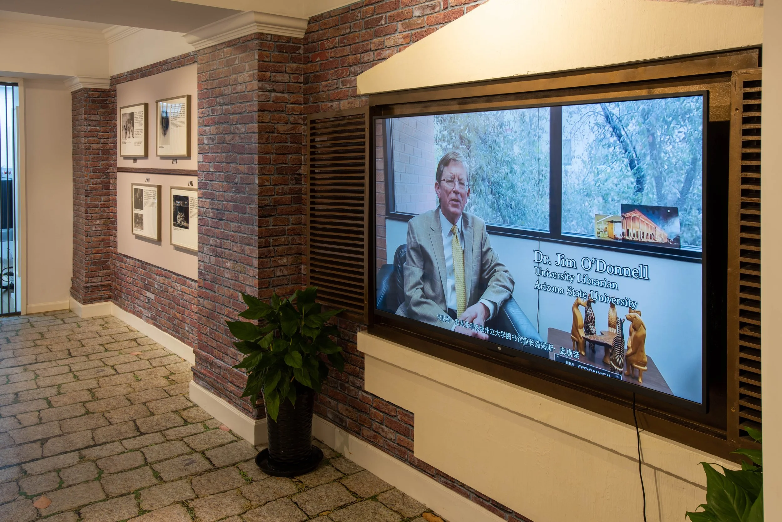 Indoor area with a brick wall and framed photos, a large digital screen displaying an interview with Dr. Jim O'Donnell, a university librarian at Arizona State University, and decorative animal figurines on a table below the screen.
