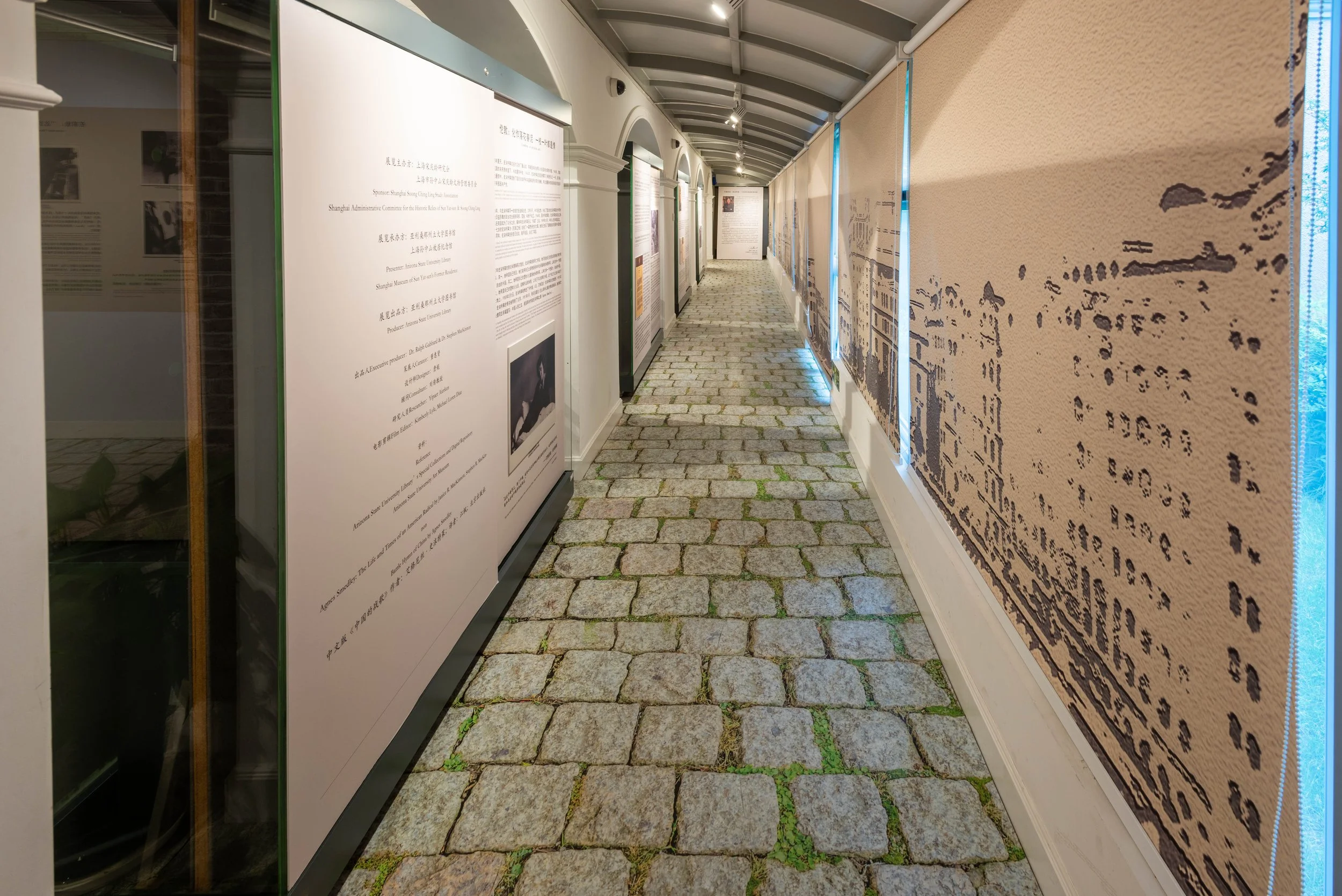 A hallway in an exhibit or museum, with informational panels on the left and a large textile or tapestry on the right, featuring a scenic or historical design, under a curved ceiling with lighting.