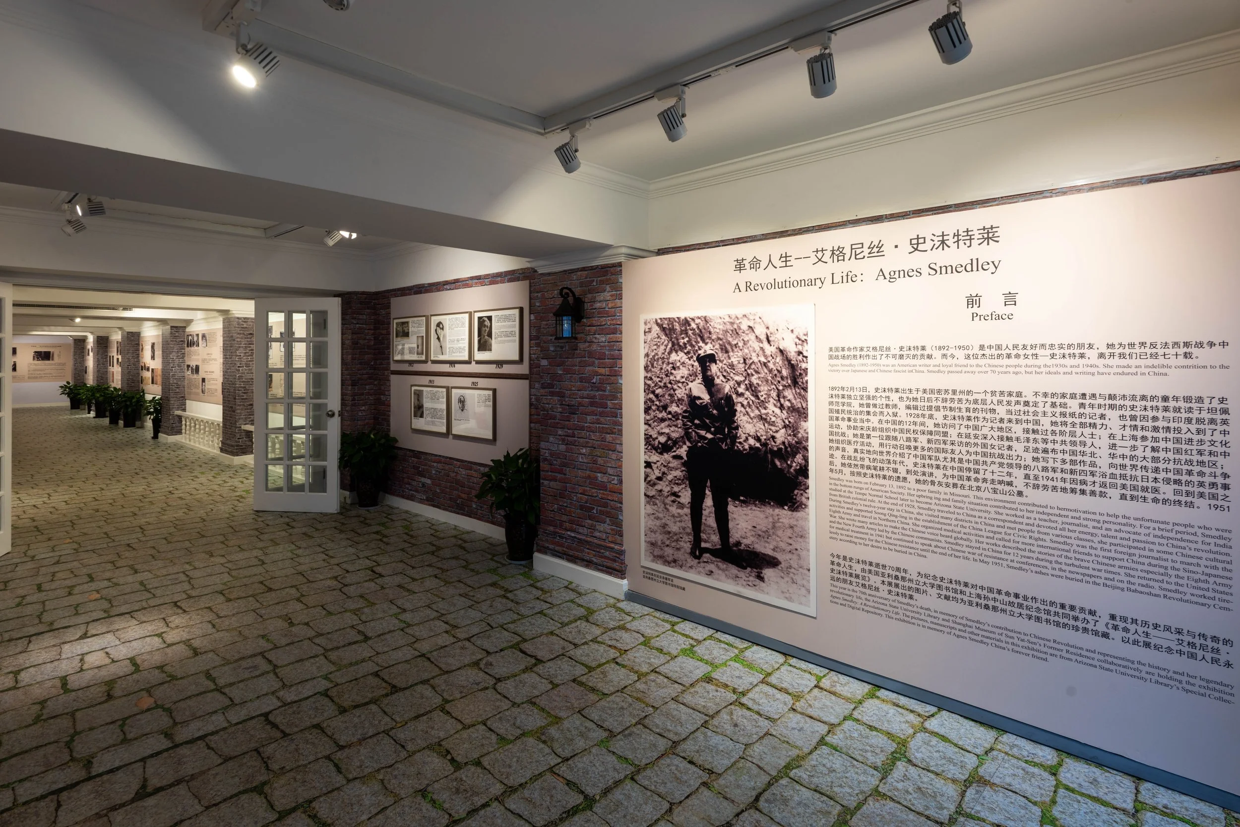 Exhibition display about revolutionary life of Agnes Smedley at a museum, featuring historical photos and informational panels on brick wall, inside a room with track lighting and cobblestone flooring.