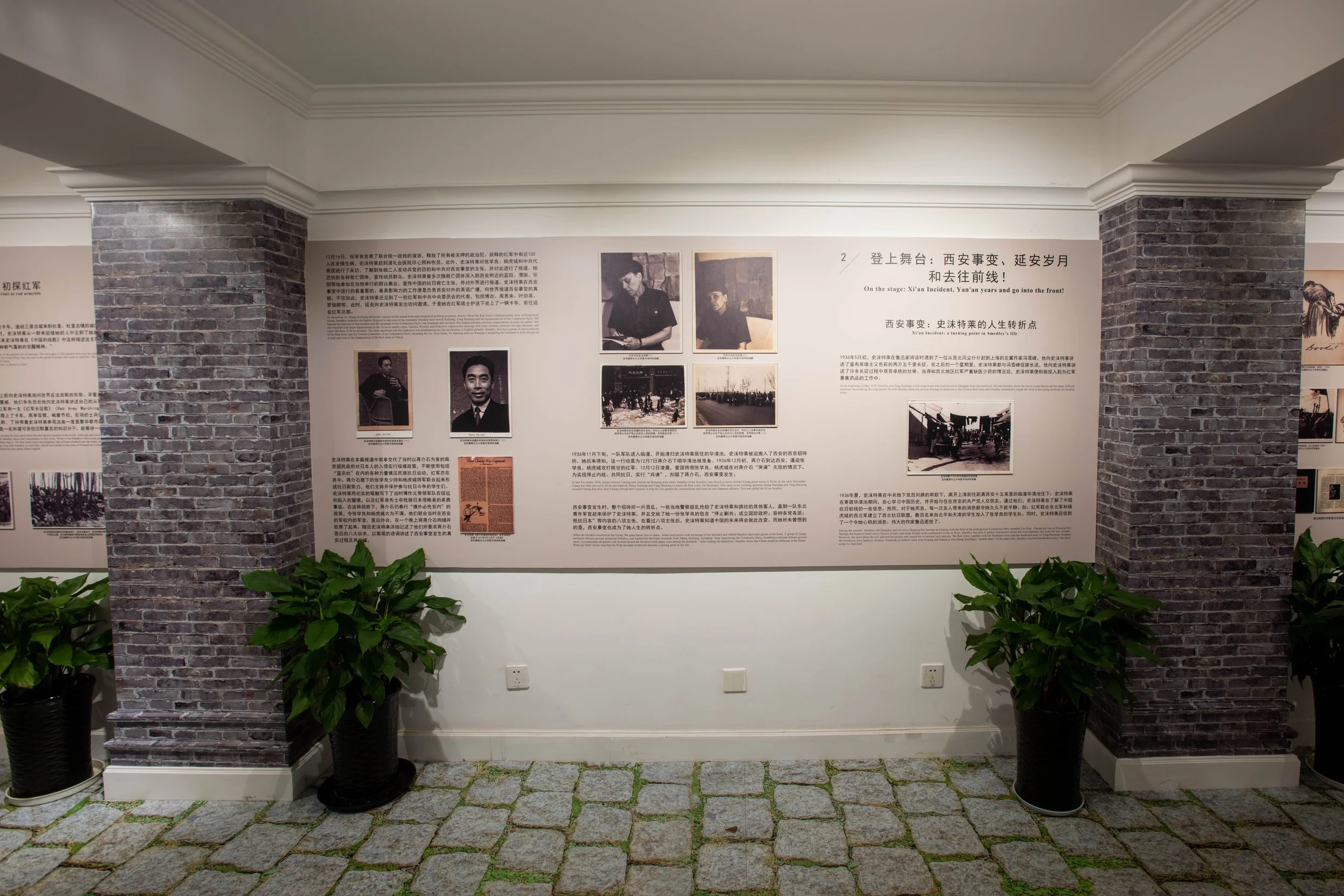 Exhibit wall with text and photographs about historical events, flanked by brick columns on white walls, with green potted plants at the bottom.