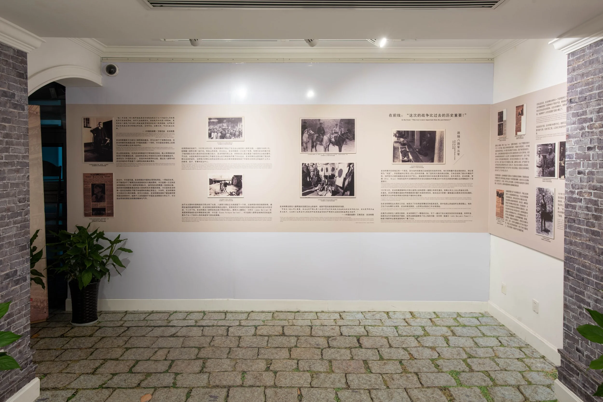 Exhibit with historical photographs, text, and Chinese characters on a wall in a gallery. Brick columns on sides, stone pavement floor, potted plant on left.