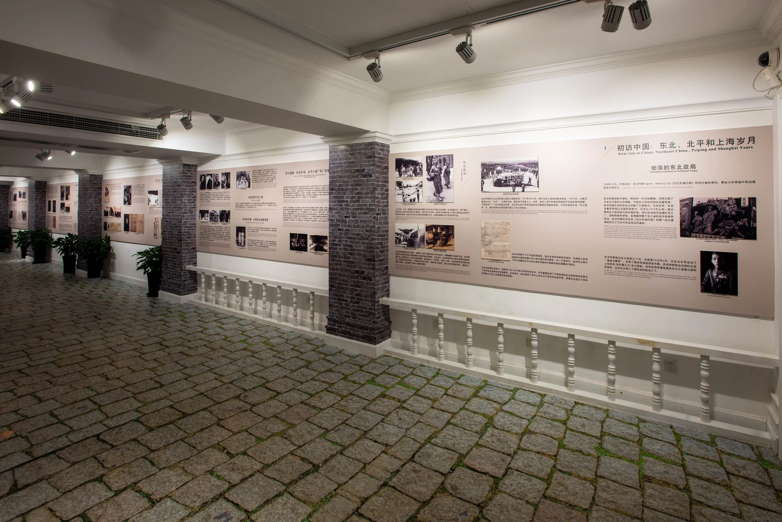An indoor exhibition space with brick columns, white walls, and framed photographs and text panels about China's history, illuminated by ceiling spotlights and decorated with potted plants along the corridor.