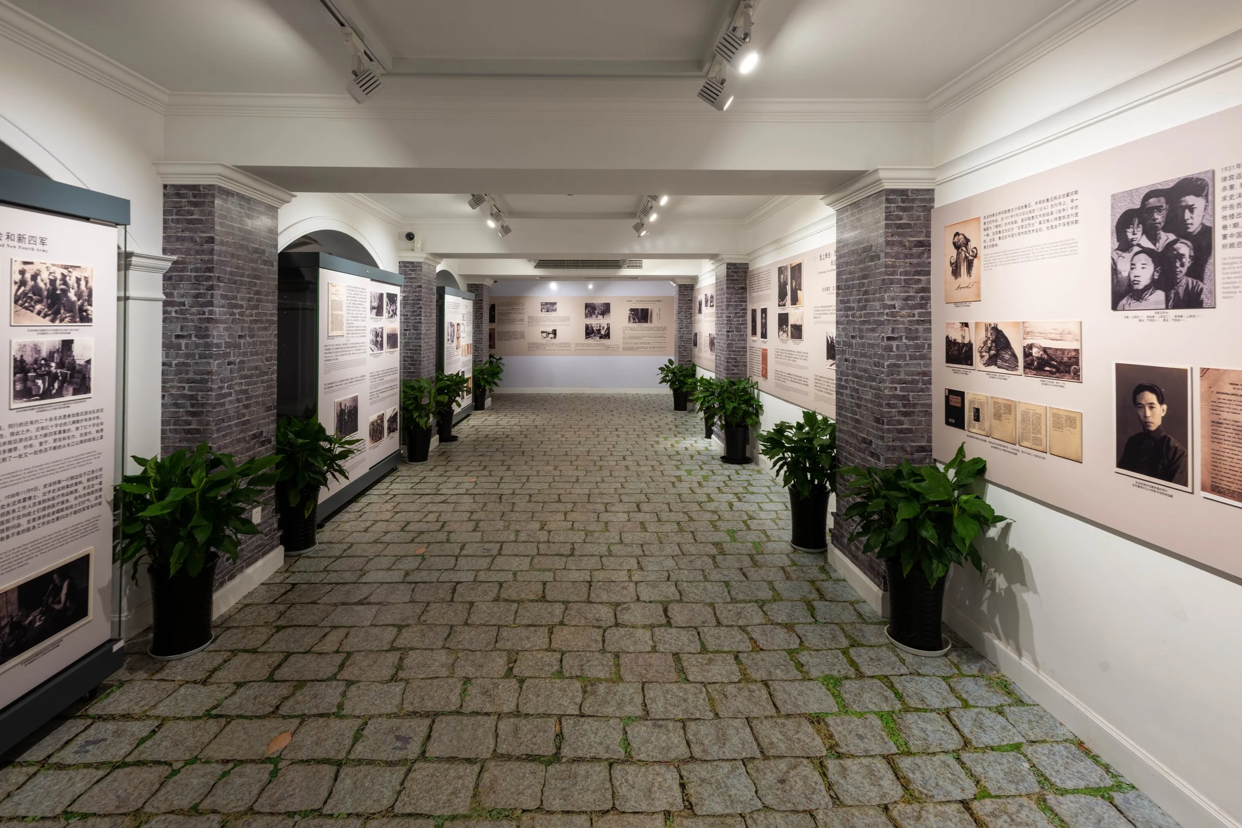 Exhibition gallery with informational panels and photographs on white walls, gray brick pillars, potted plants, and cobblestone floor.